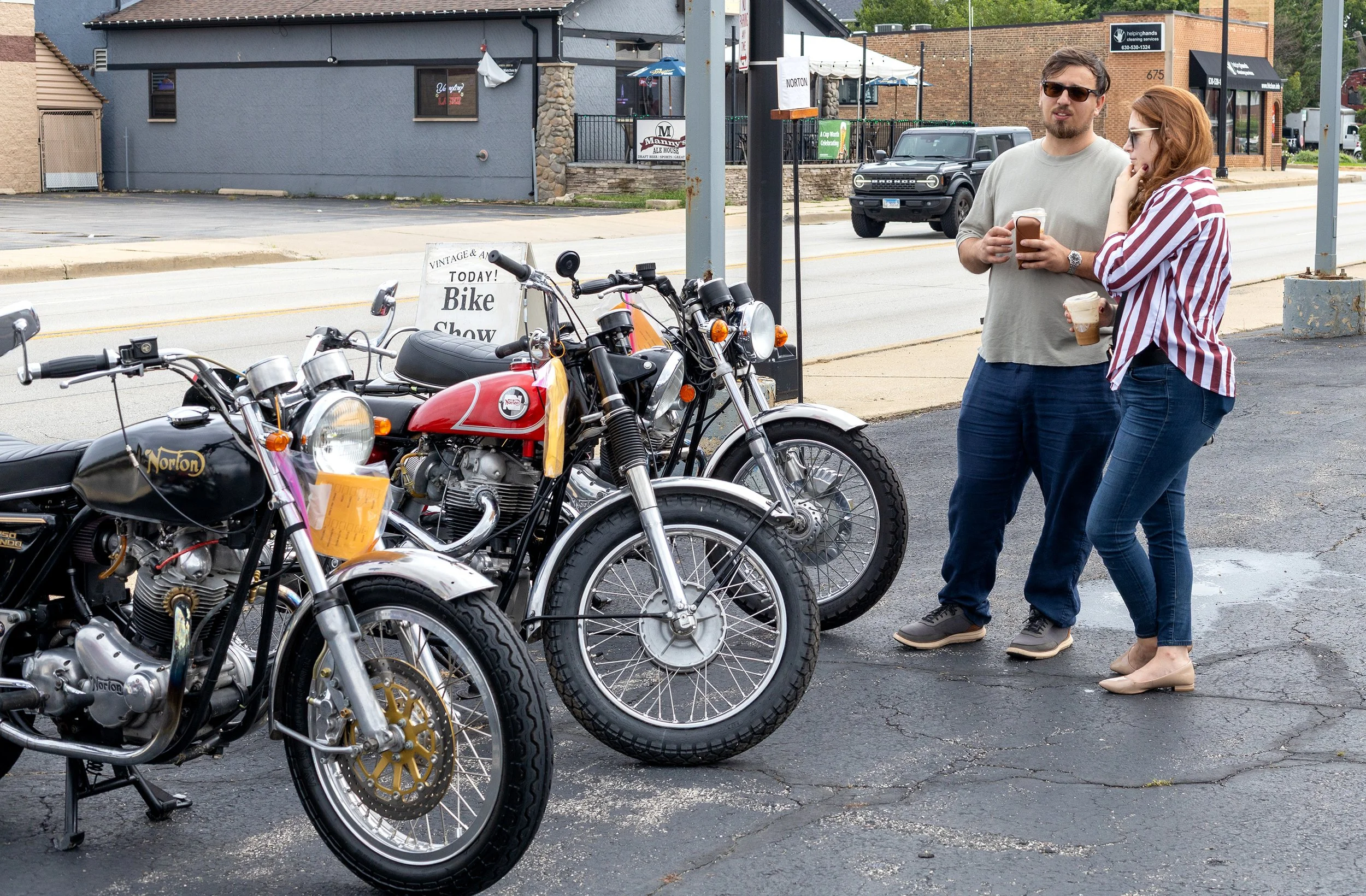 Two people, a man with glasses and a woman with red hair, standing on a sidewalk with motorcycles in front of them, holding coffee cups, during a bike show event.