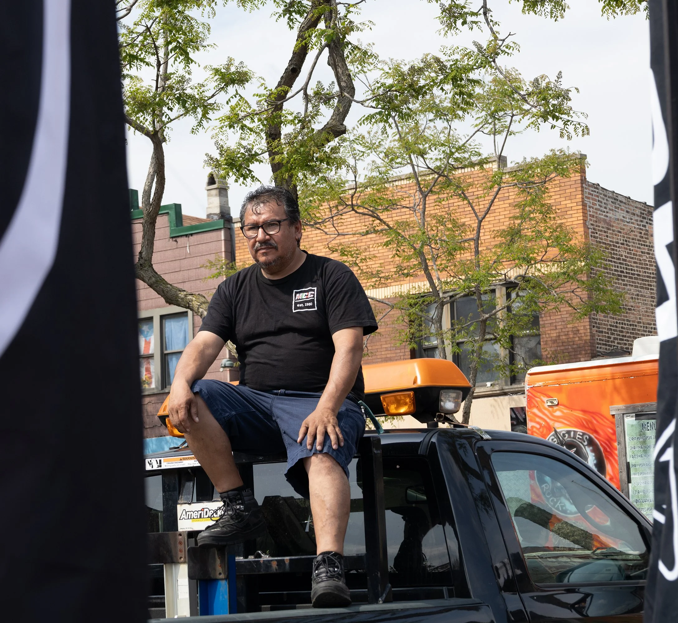 A man with glasses and a black T-shirt sits on a black vehicle, seen through a gap between objects. Behind him are buildings and trees in an outdoor urban setting.
