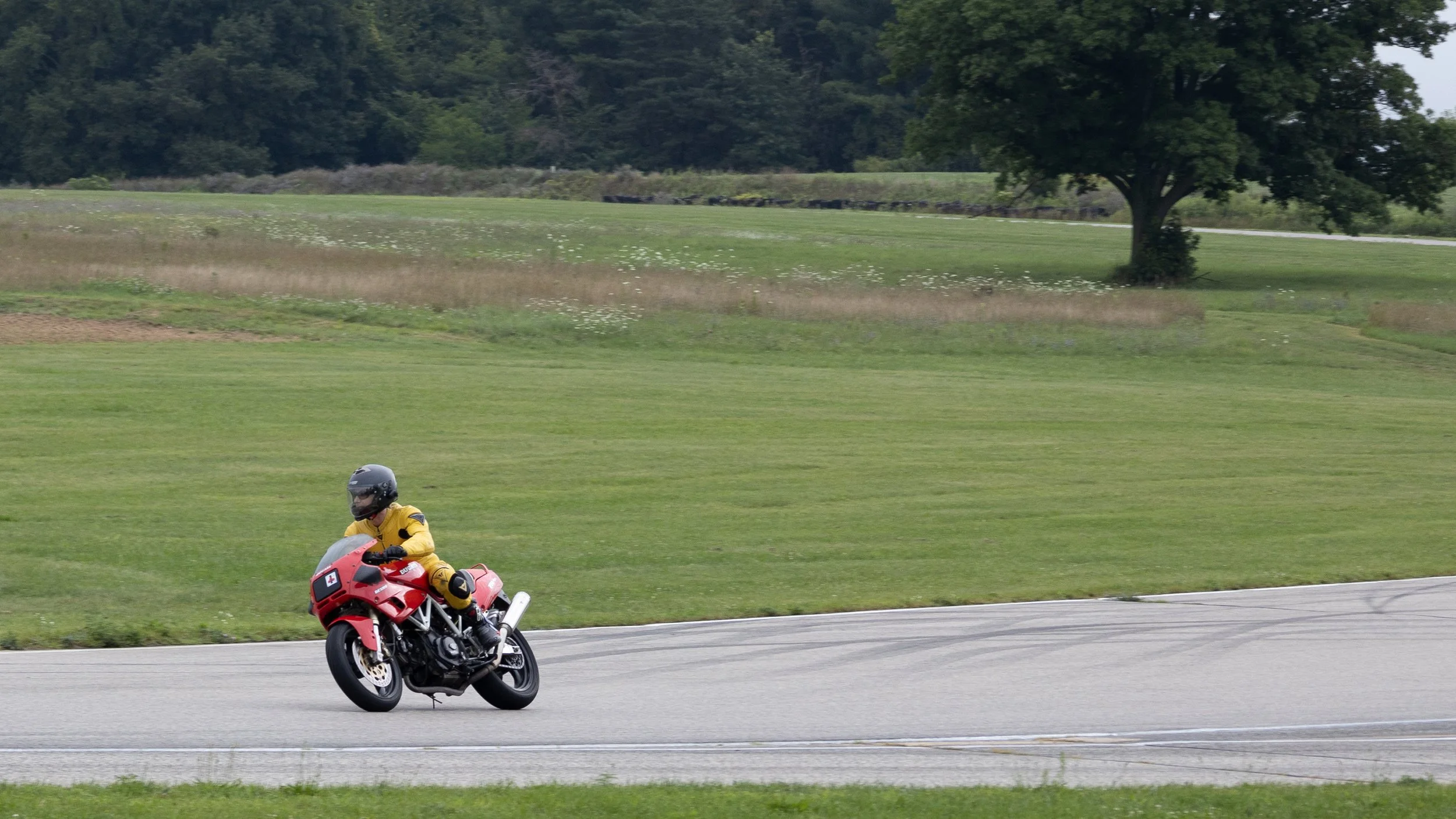 A person riding a red motorcycle on a paved path, wearing a yellow jacket and helmet, with green grassy fields and trees in the background.