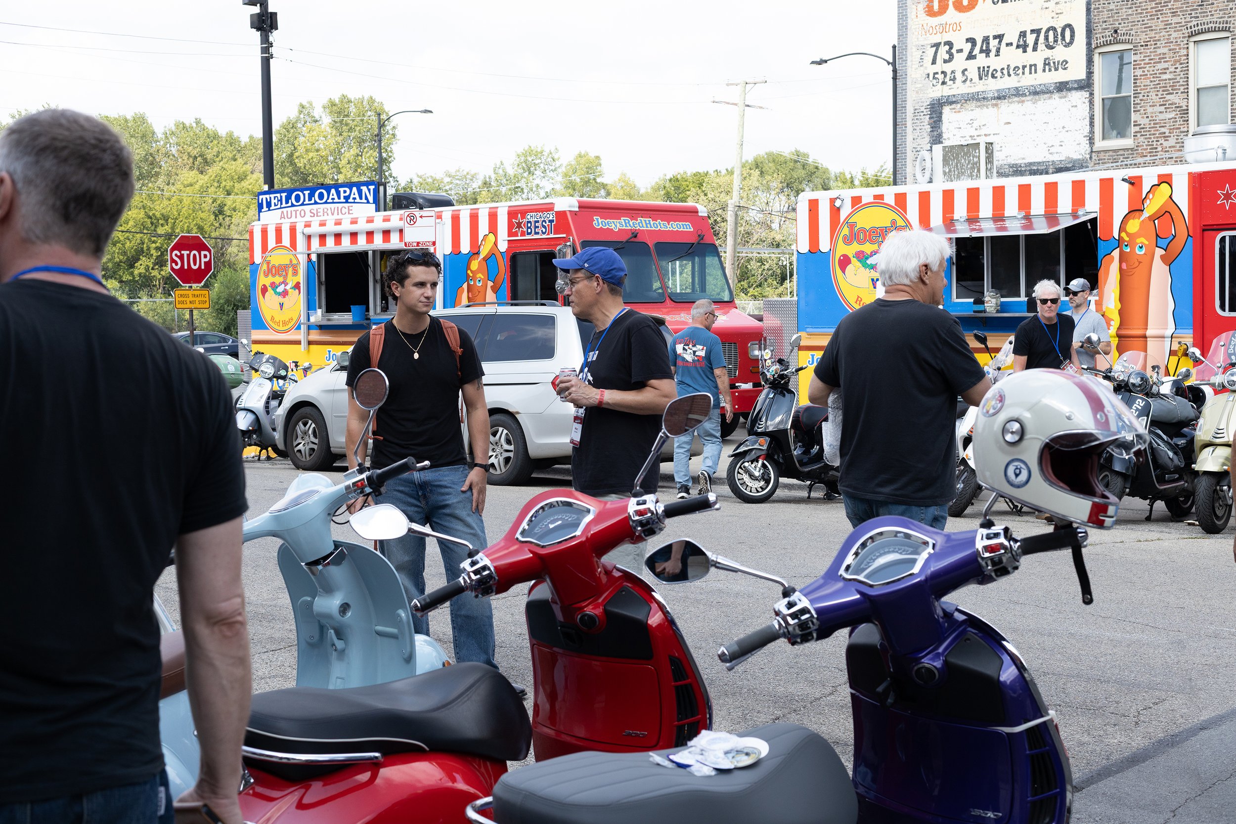 A street scene with several parked scooters and motorcycles, with people walking and talking. Food trucks with bright, colorful designs are in the background, along with a large brick building and trees.