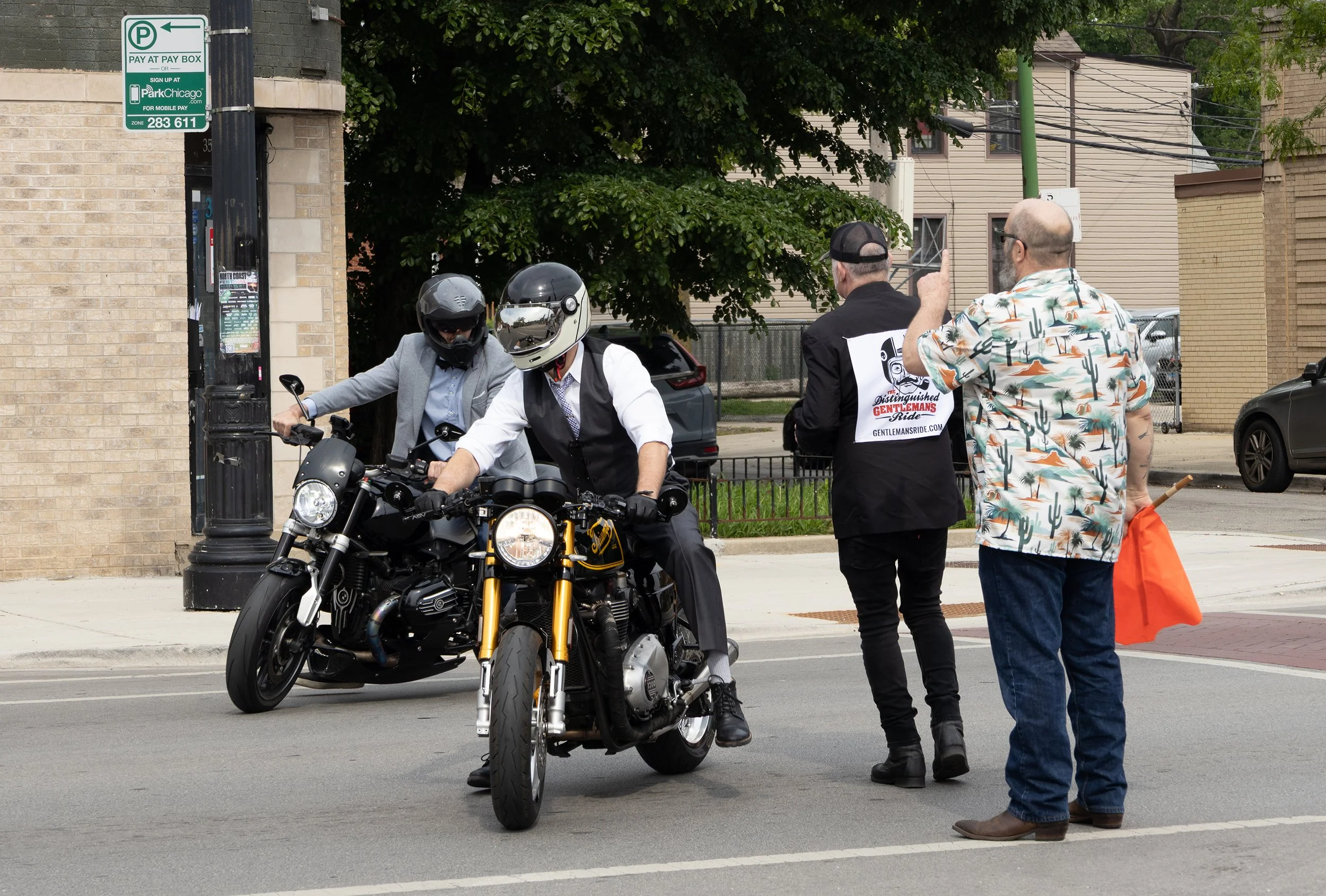 Two men on a motorcycle stop at a street corner talking to a woman and a man. The woman is wearing a hat with a black vest that has a logo and text, and the man is wearing a tropical print shirt. The man on the motorcycle is wearing a grey helmet and