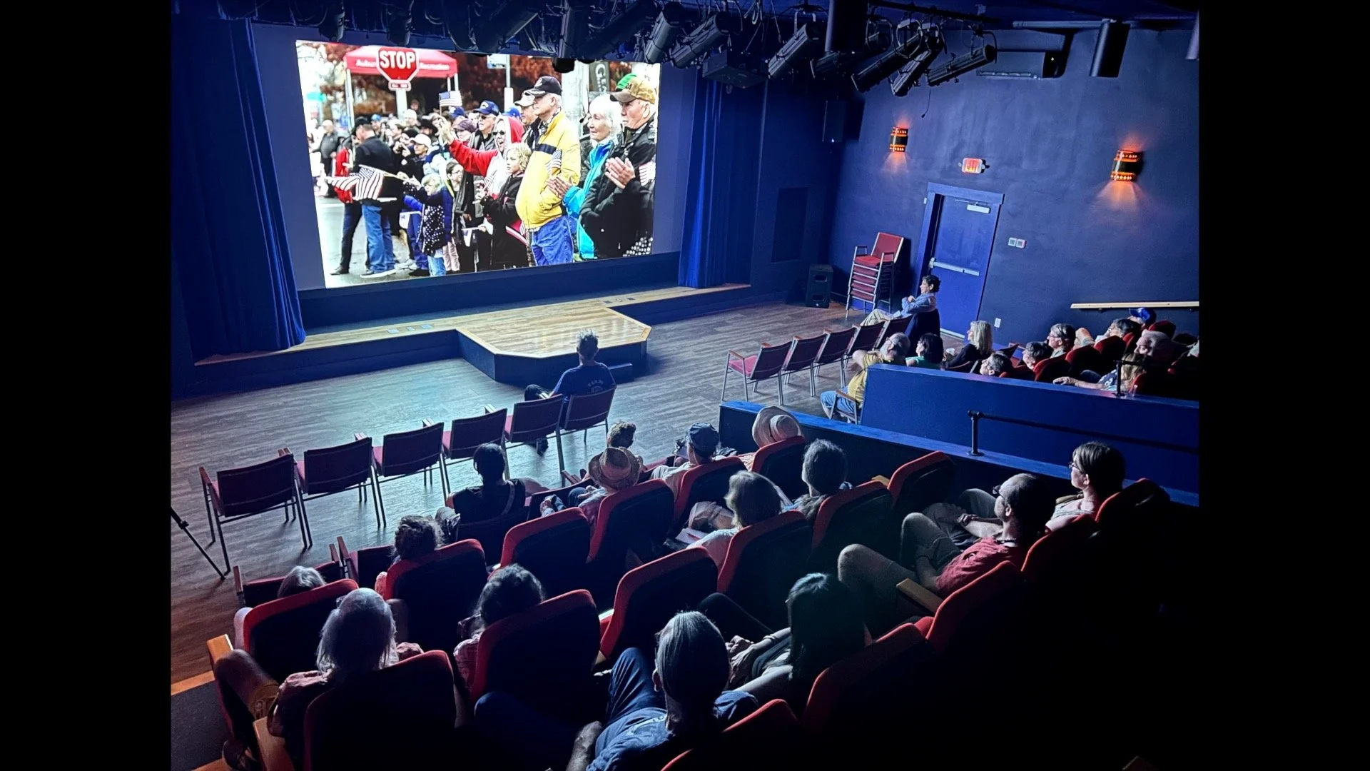 Small theater with seats filled, everyone's watching the film that shows people at a parade waving flags