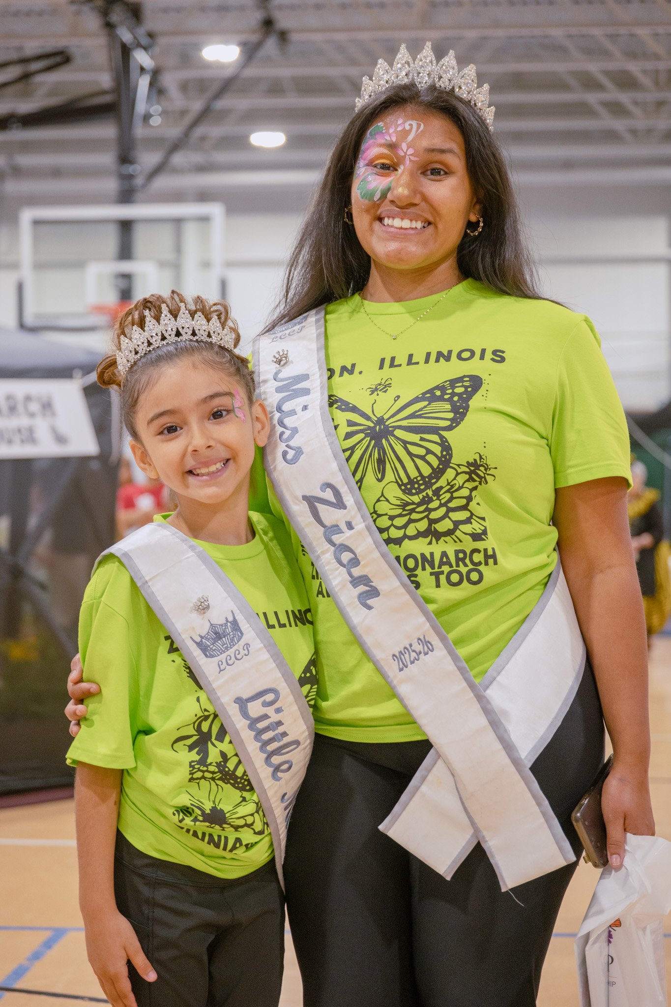 Two women wearing matching white “Zion, Illinois – Monarch & Bees Too Festival” T-shirts with a colorful butterfly graphic stand arm in arm on a grassy lawn, smiling at the camera.