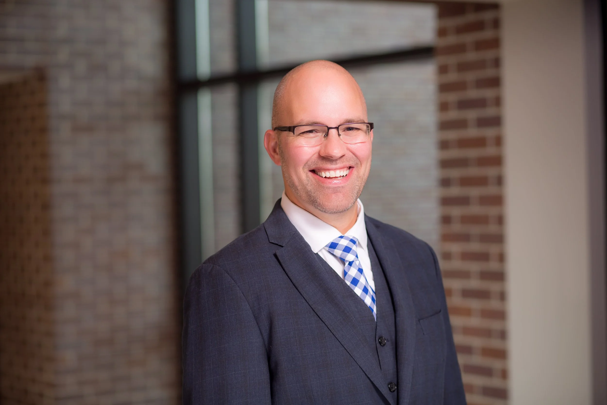 A smiling man in a business suit and glasses standing indoors near a brick and glass wall.