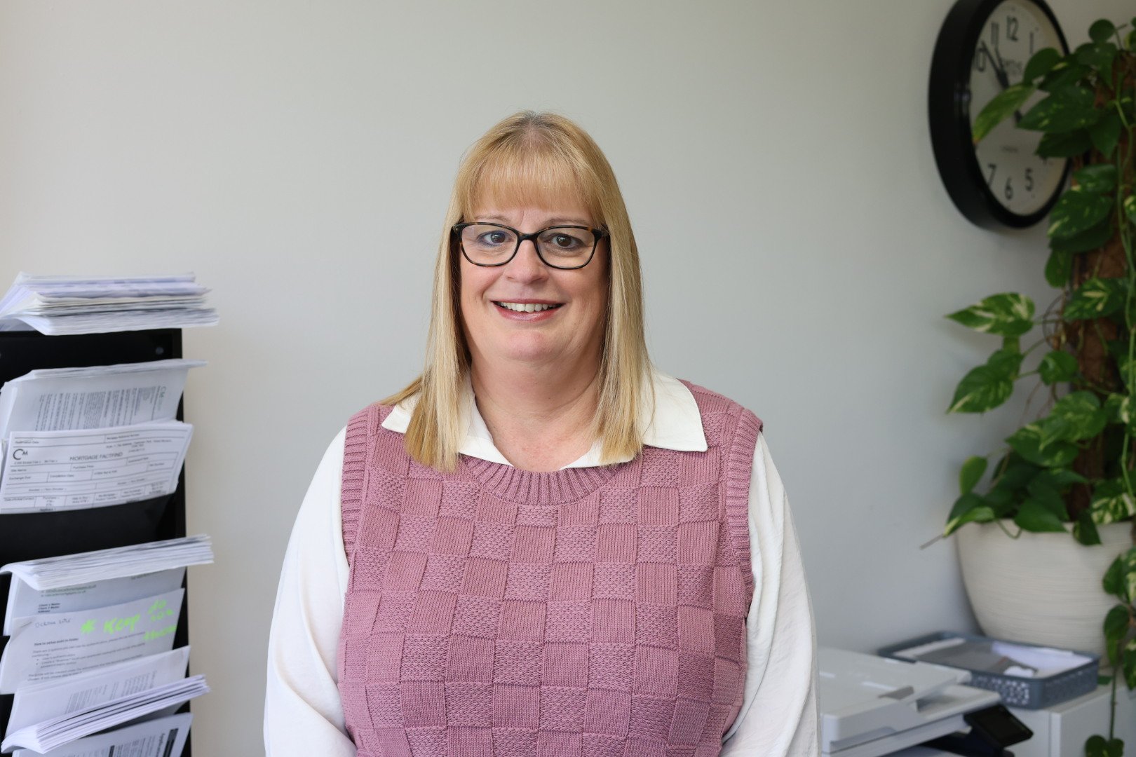 A woman with shoulder-length blond hair, wearing glasses and a cream-colored top with lace details, smiling against a plain white background.