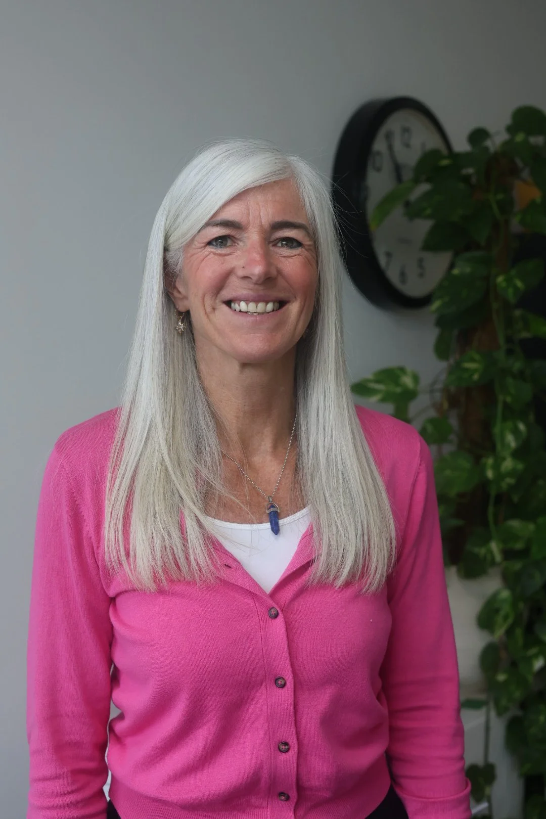 A woman with shoulder-length gray hair wearing a colorful geometric patterned blouse, earrings, and a necklace, standing against a plain white wall, smiling at the camera.