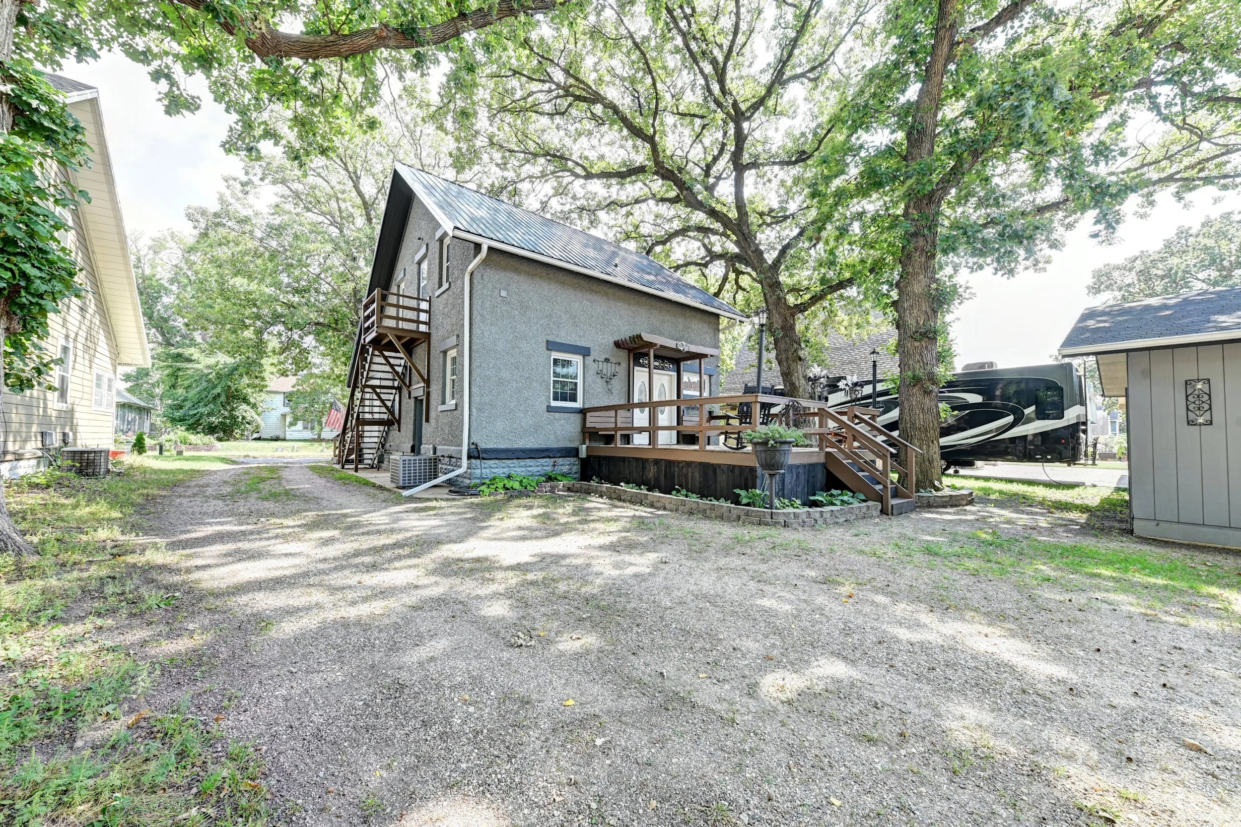 A residential backyard with a gravel ground, a two-story house with a staircase, large trees providing shade, and a person RV parked nearby.