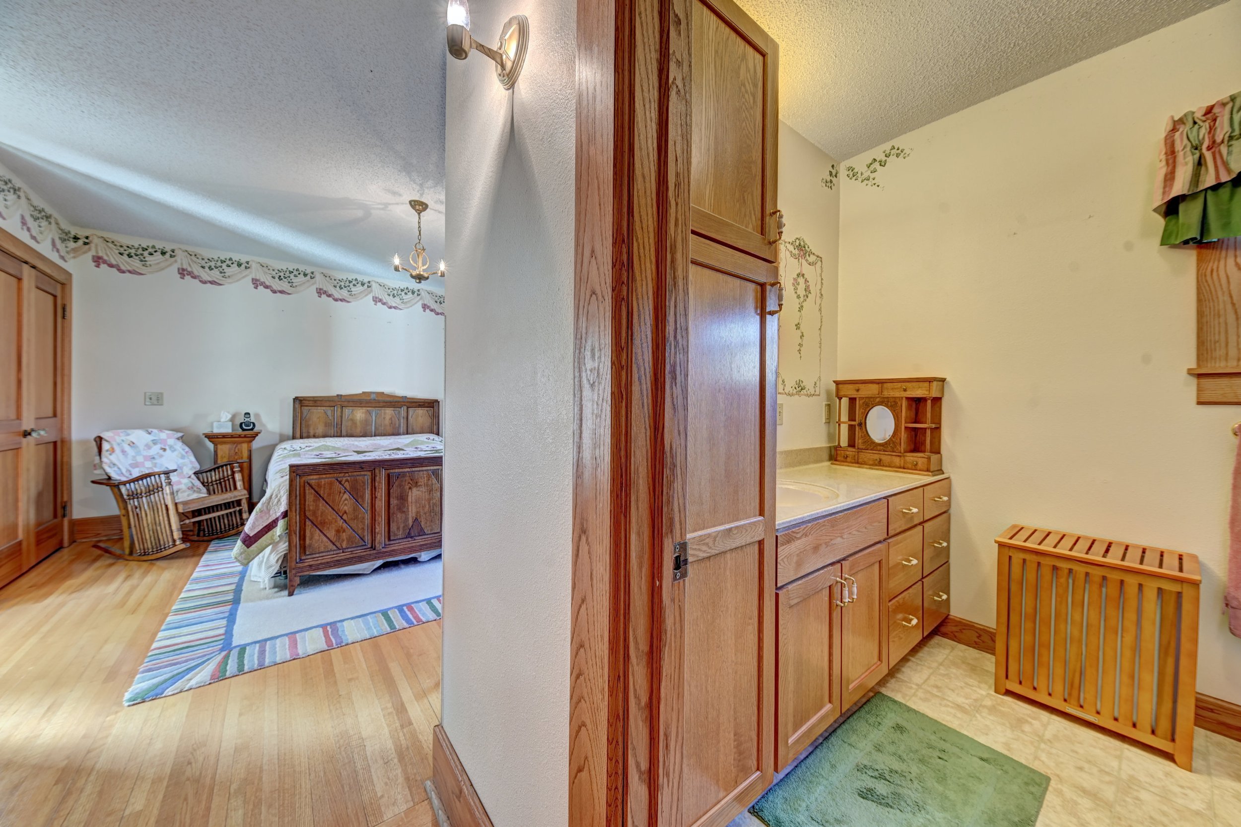 View from the hallway into a bedroom with a wooden bed, a quilt, a wooden nightstand, and a floral border on the wall. The hallway has wooden cabinets and a small green rug near a wooden baby crib.