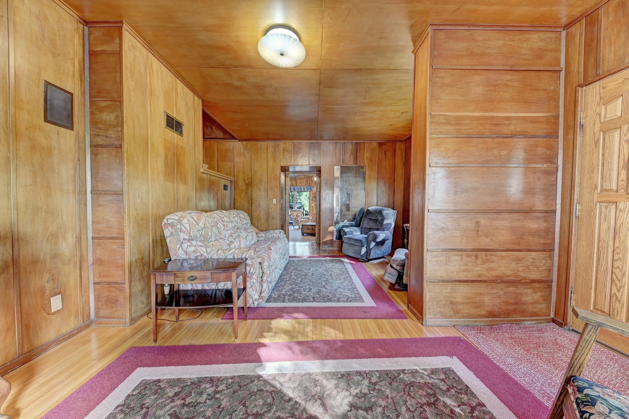 Living room with wood-paneled walls, hardwood floor, area rug, and furniture including a floral sofa, armchairs, and a wooden side table.
