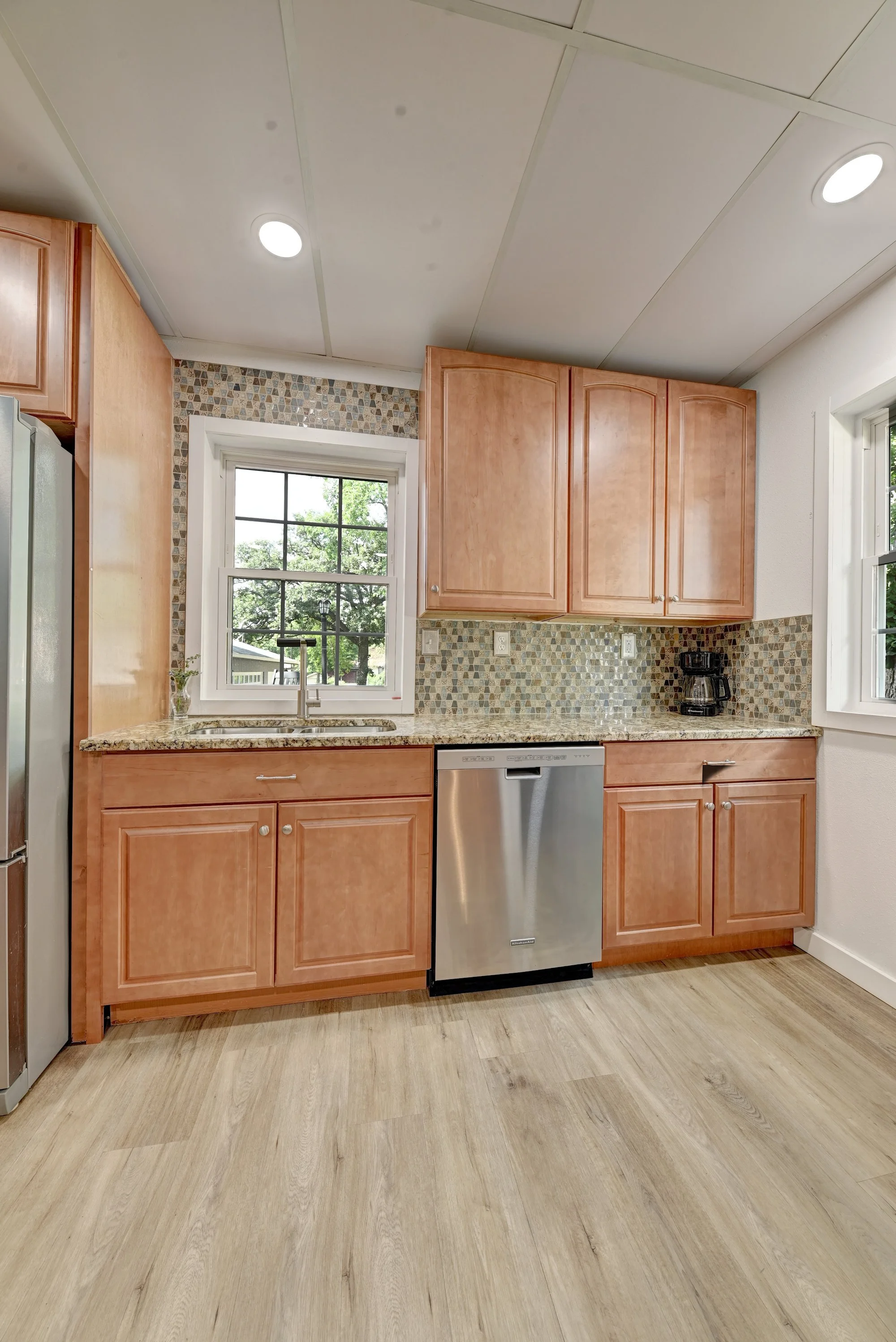 A kitchen with light wooden cabinetry, a granite countertop, a mosaic tile backsplash, a window above the sink, a coffee maker, and a dishwasher.