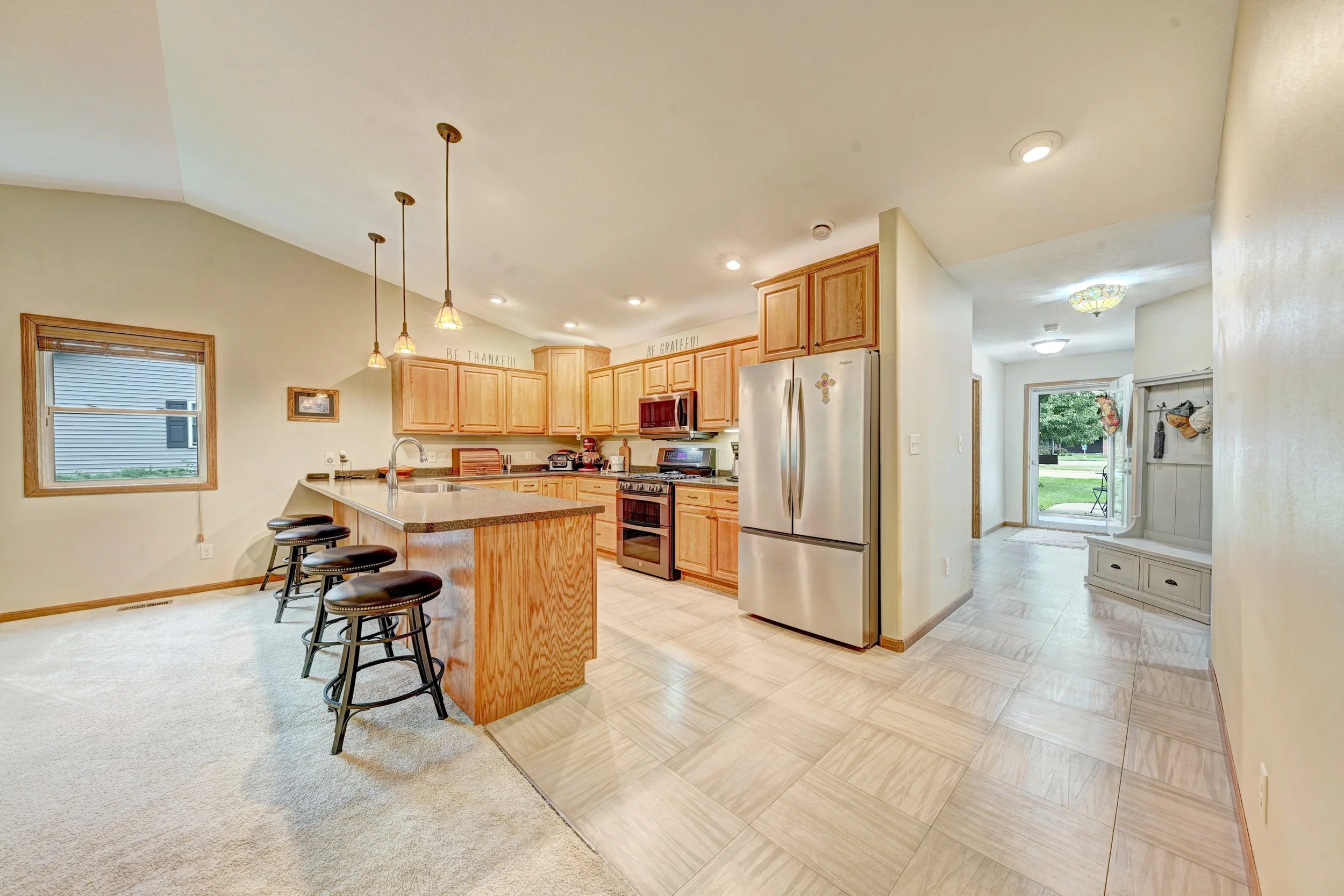 Interior view of a kitchen with light wood cabinets, a stainless steel refrigerator, and a kitchen island with three black stools. There are pendant lights hanging above the island and a window with a view outside. The kitchen is connected to a room 