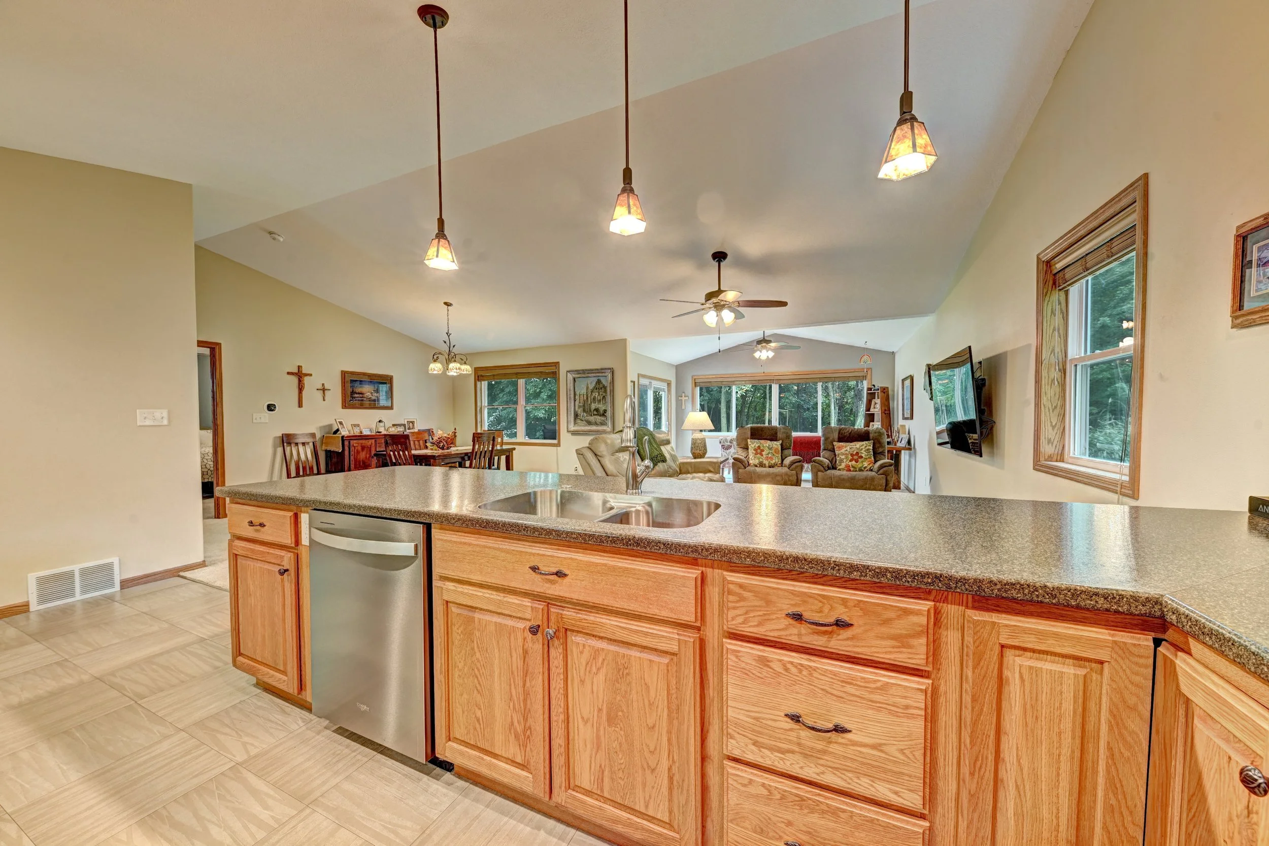 Open-concept kitchen and living room with pendant lights, ceiling fans, residential windows, a dining area, and a flat-screen TV on the wall.