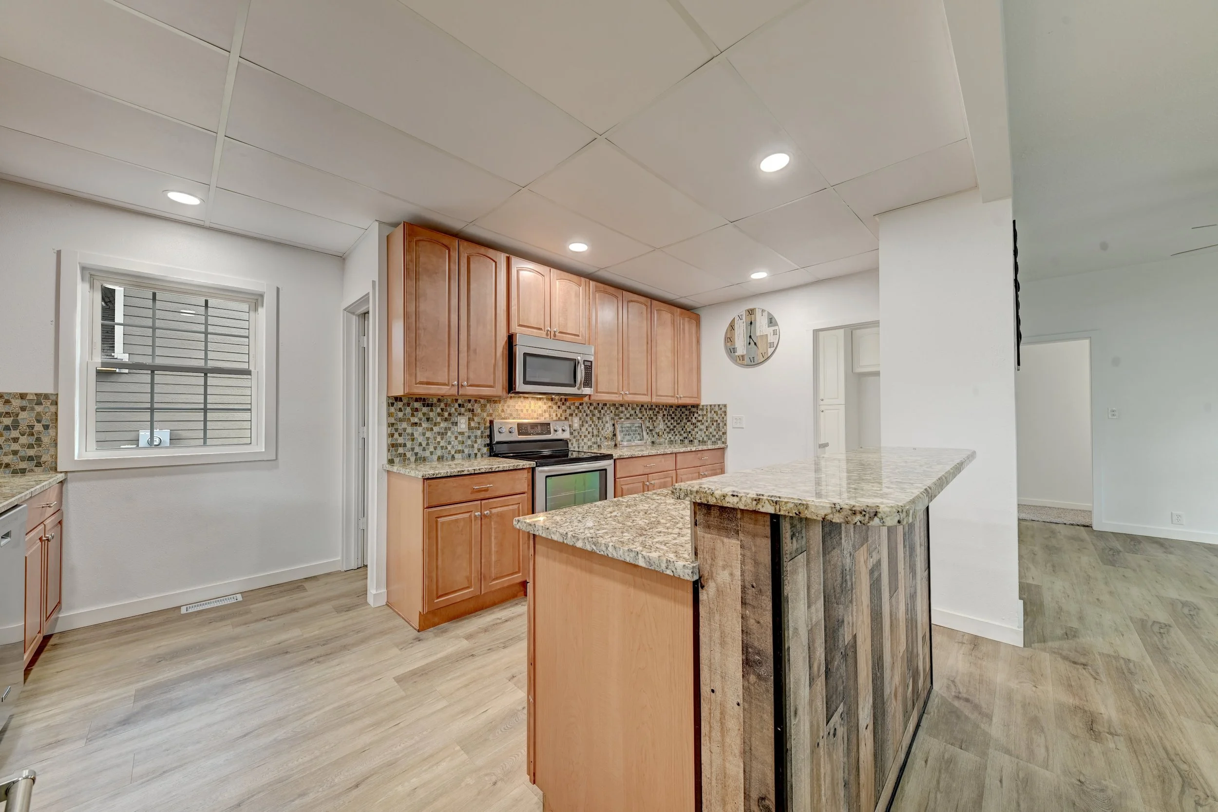 A kitchen with wooden cabinets, granite countertops, recessed lighting, a microwave, and a clock on the wall.