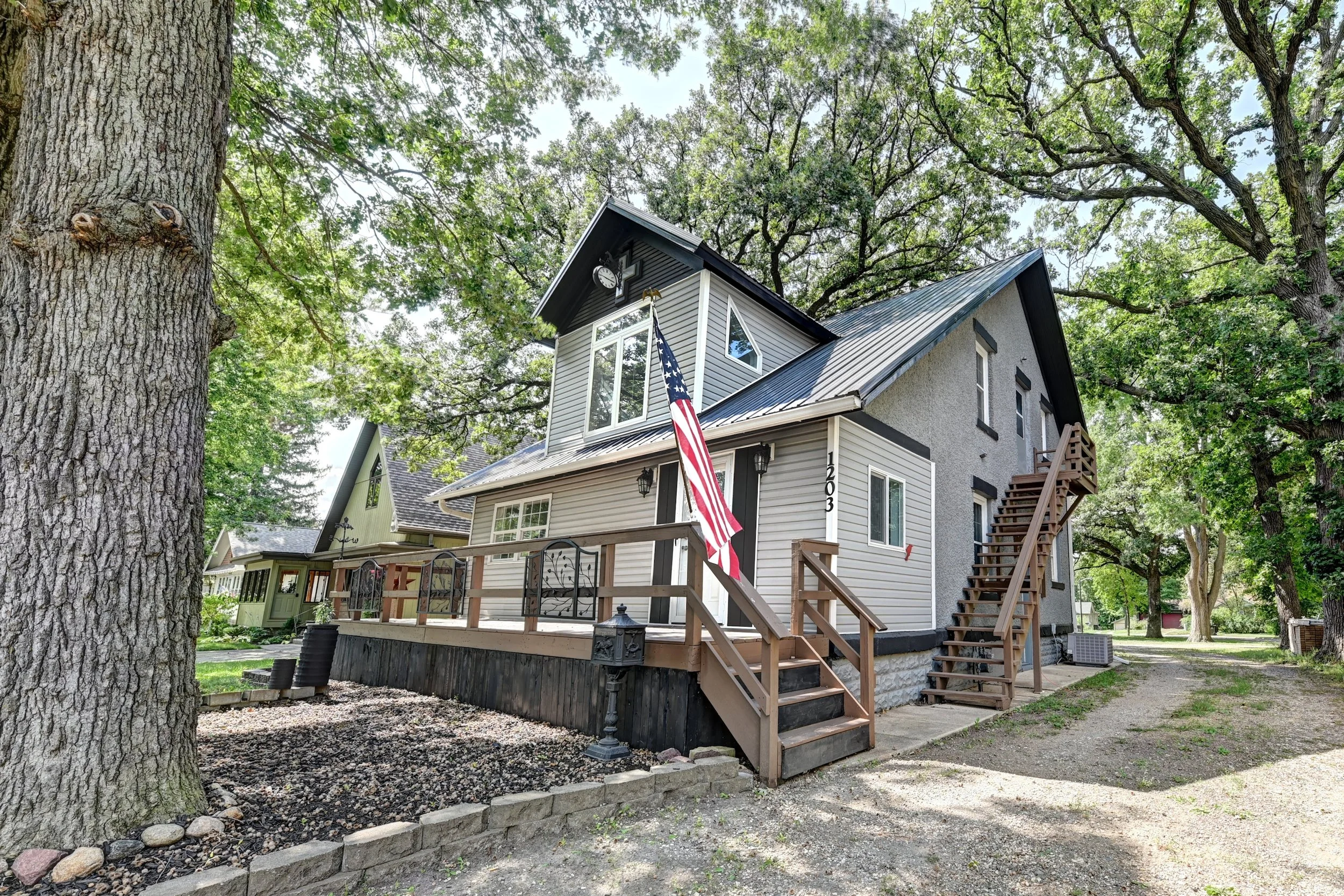 A two-story house with gray and white exterior, a front porch with stairs, an American flag hanging, and a large tree in the yard, surrounded by other trees under a partly cloudy sky.