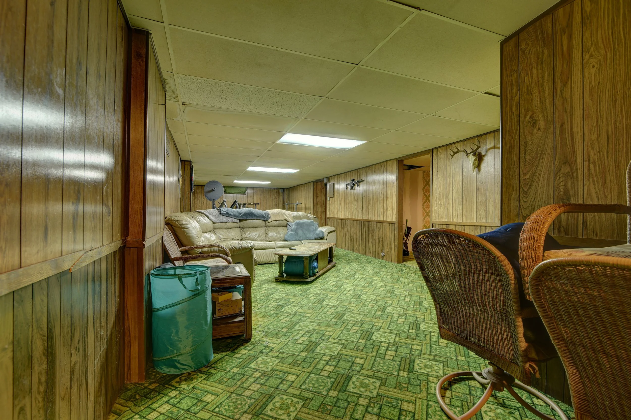 A basement room with wood-paneled walls, a beige leather sofa, wicker chairs, and a small wooden table on a green patterned carpet. The ceiling has white panels, with some lights and a ceiling vent. In the background, there are mounted guns and a dee