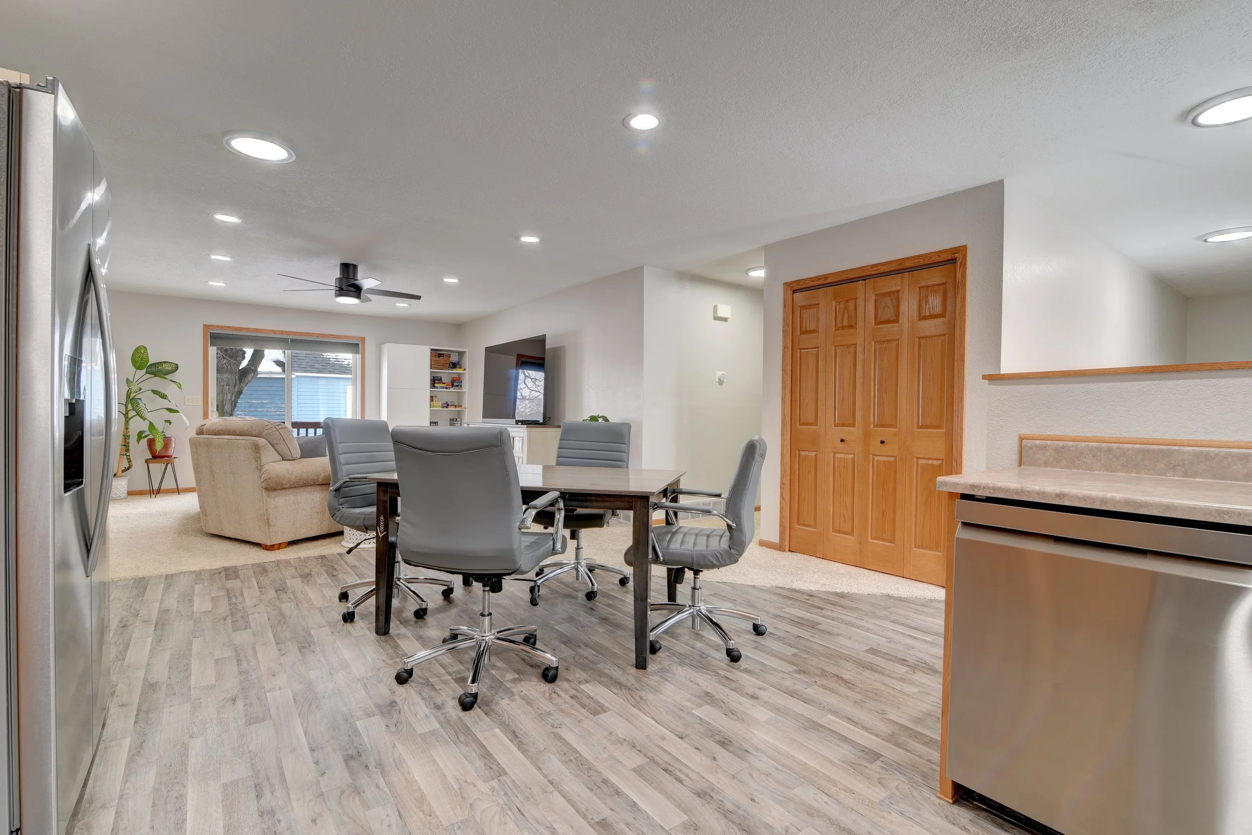 Open-concept living room and dining area with hardwood floors, cream-colored walls, and wood accents. Features a dining table with five gray chairs, a beige sofa, a TV on a white stand, and large windows with a view of the outdoors.