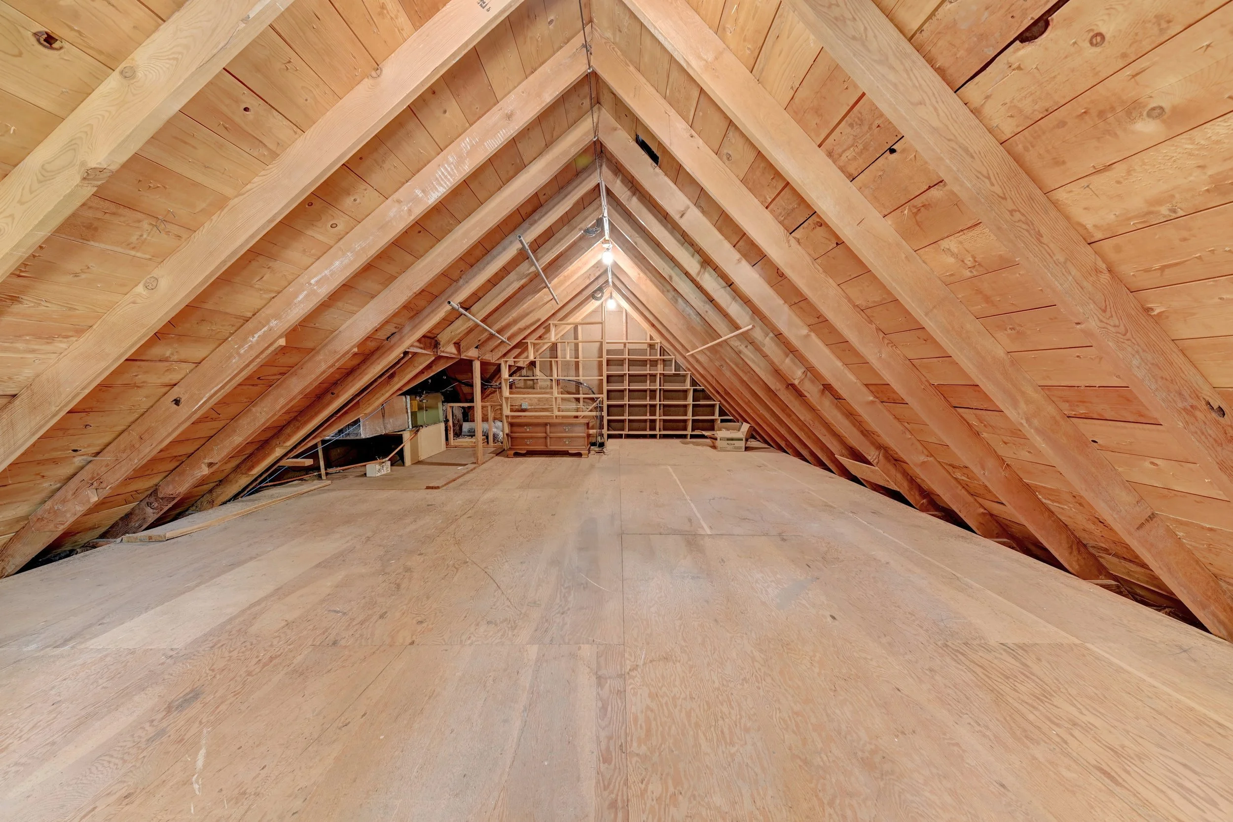 Attic space with unfinished wooden roof framing and walls, construction materials, and a hanging light bulb.