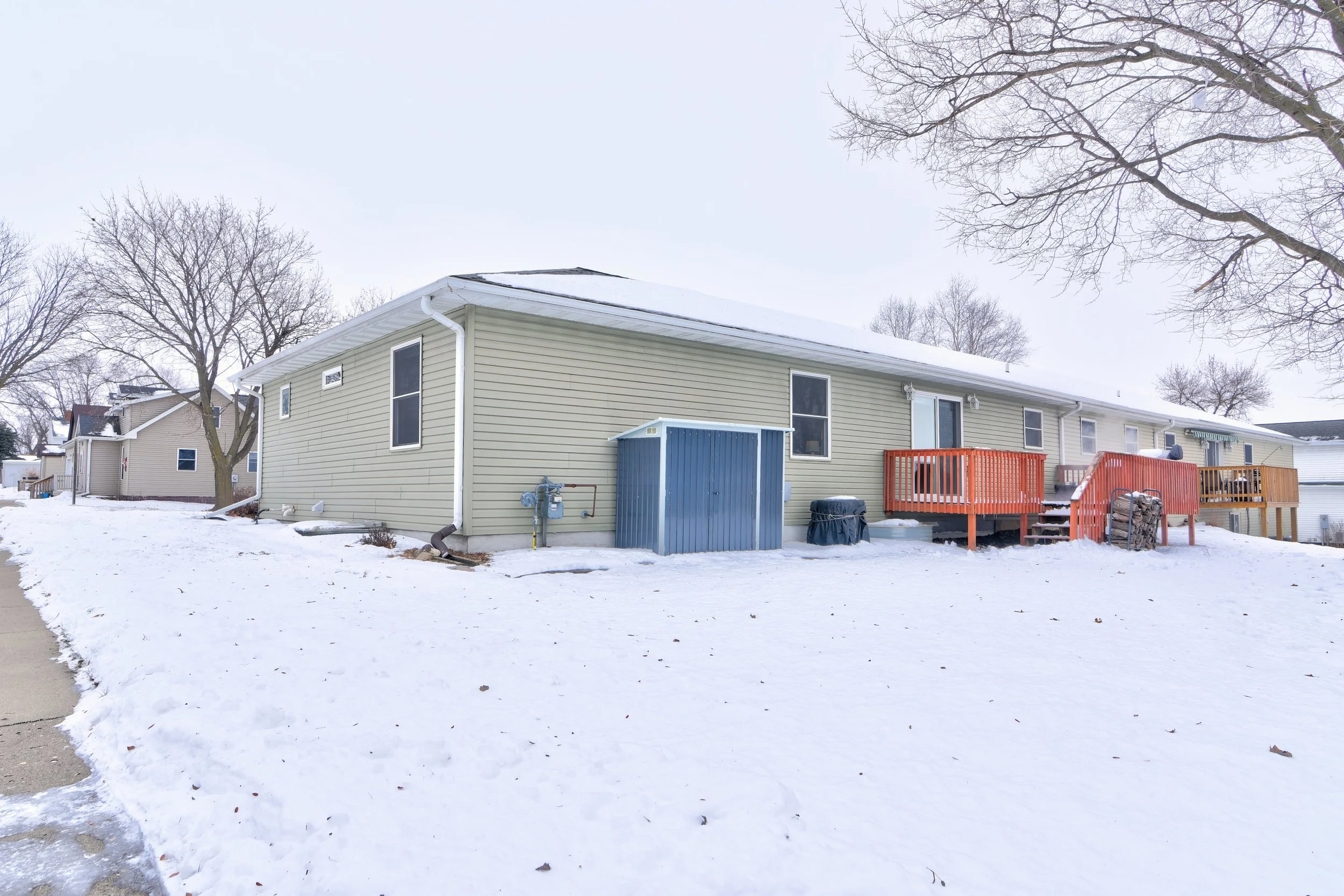 Backyard view of a beige house with orange and blue decks, snow covering the ground, leafless trees, and neighboring houses in the background.