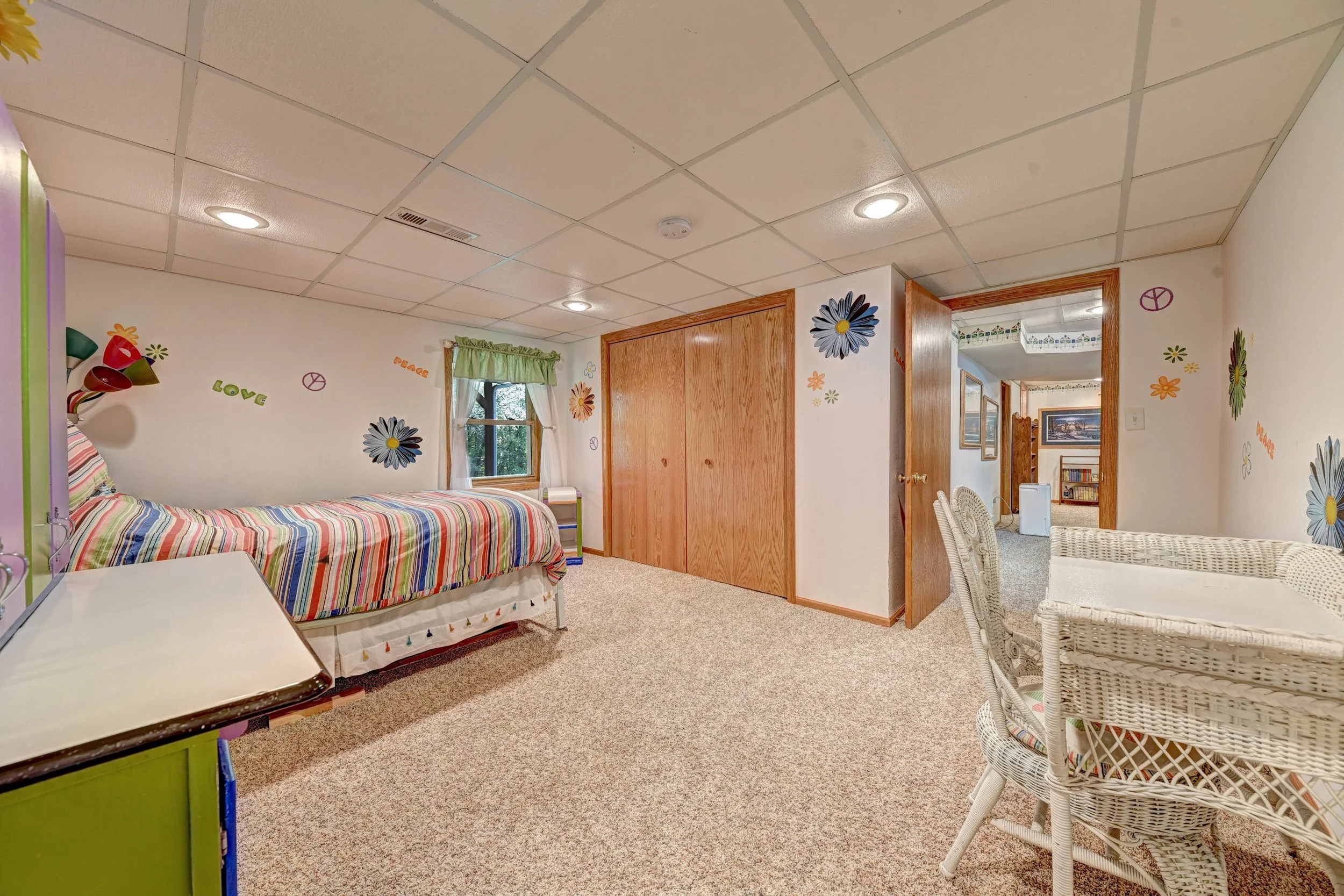 A colorful room with a single bed covered in a striped blanket, decorated with flowers and peace signs on the cream-colored walls. There is a window with green curtains, a white wicker chair, and a wooden closet with double doors.