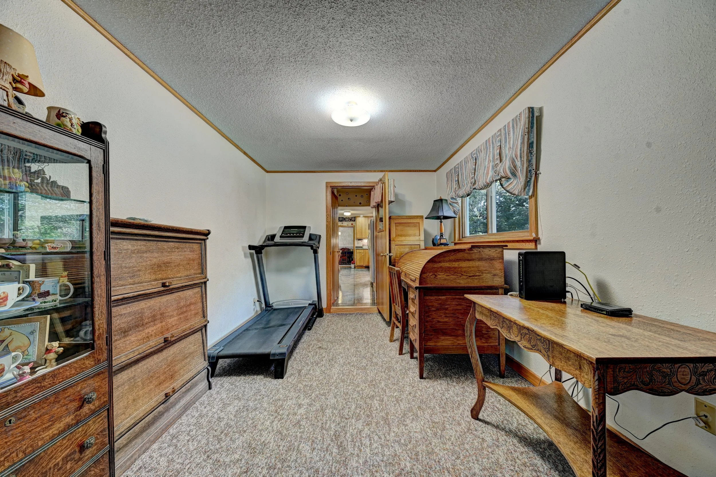 A room with a treadmill, a wooden display cabinet with collectibles, a wooden desk, a window with a striped valance, and a lamp, in a home interior.