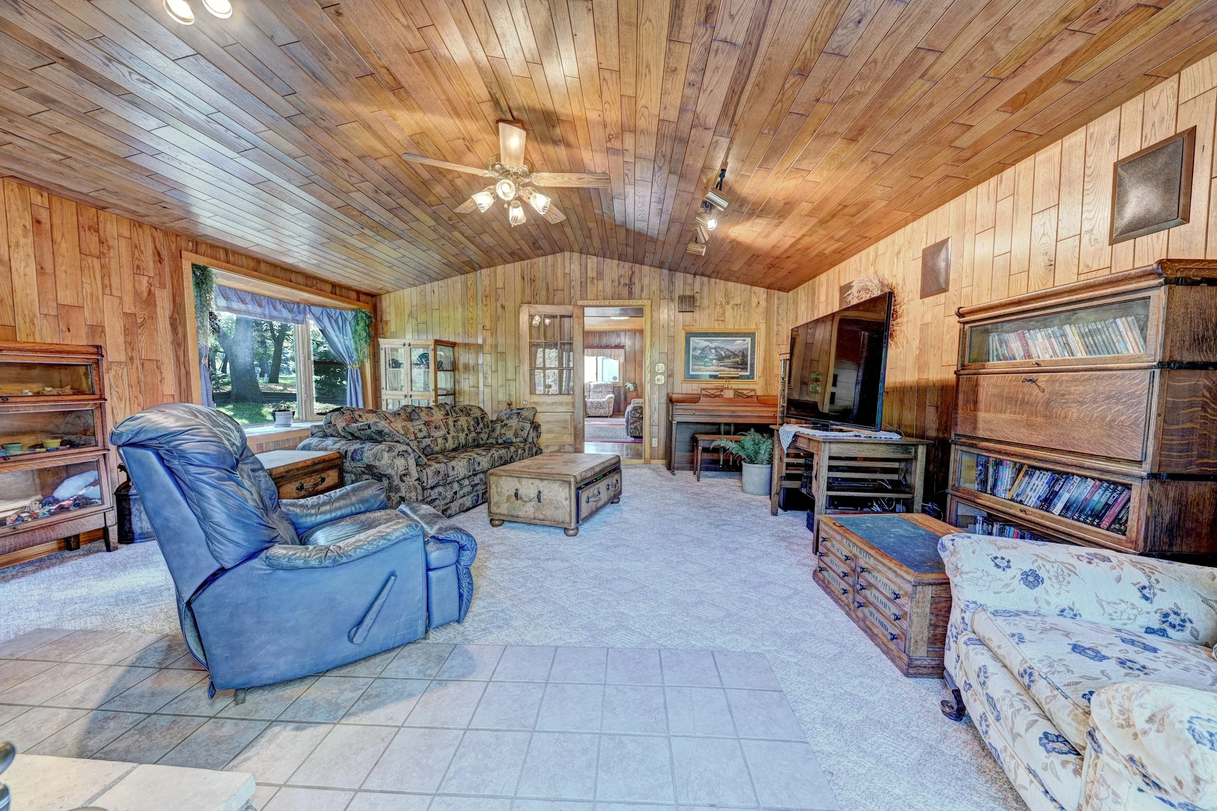 Living room with wood-paneled walls and ceiling, furnished with a floral sofa, leather chairs, a TV, bookshelves, and a window with curtains showing trees outside.