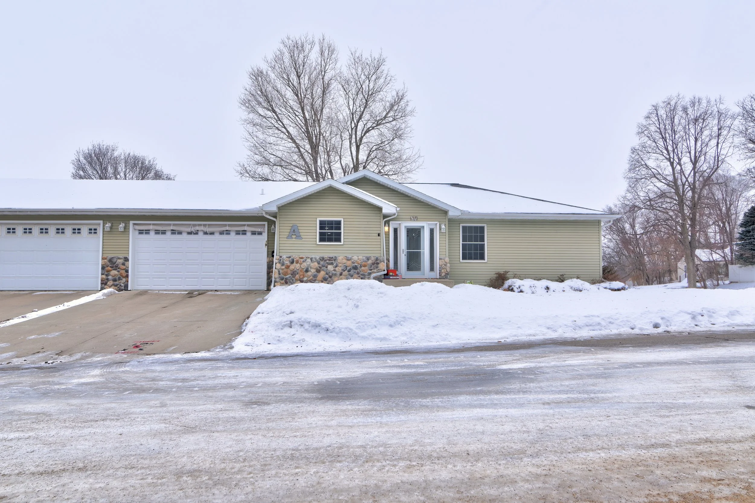 A house covered in snow with a driveway, trees, and overcast sky in the background.