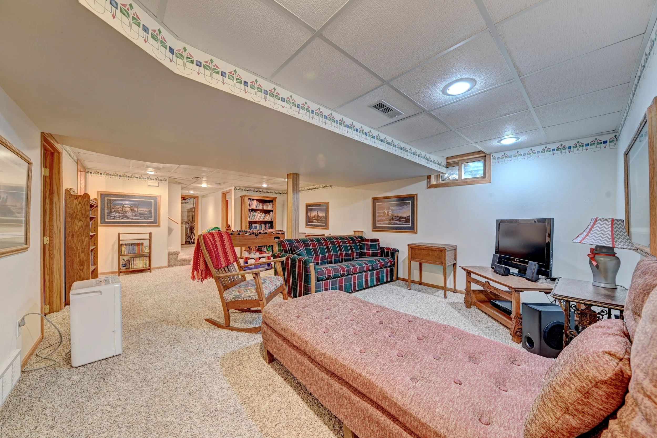 A cozy living room with beige carpet and white walls decorated with framed landscape paintings and a patterned border near the ceiling. There is a pink upholstered bench in the foreground, a plaid couch to the right, and a wooden coffee table with a 