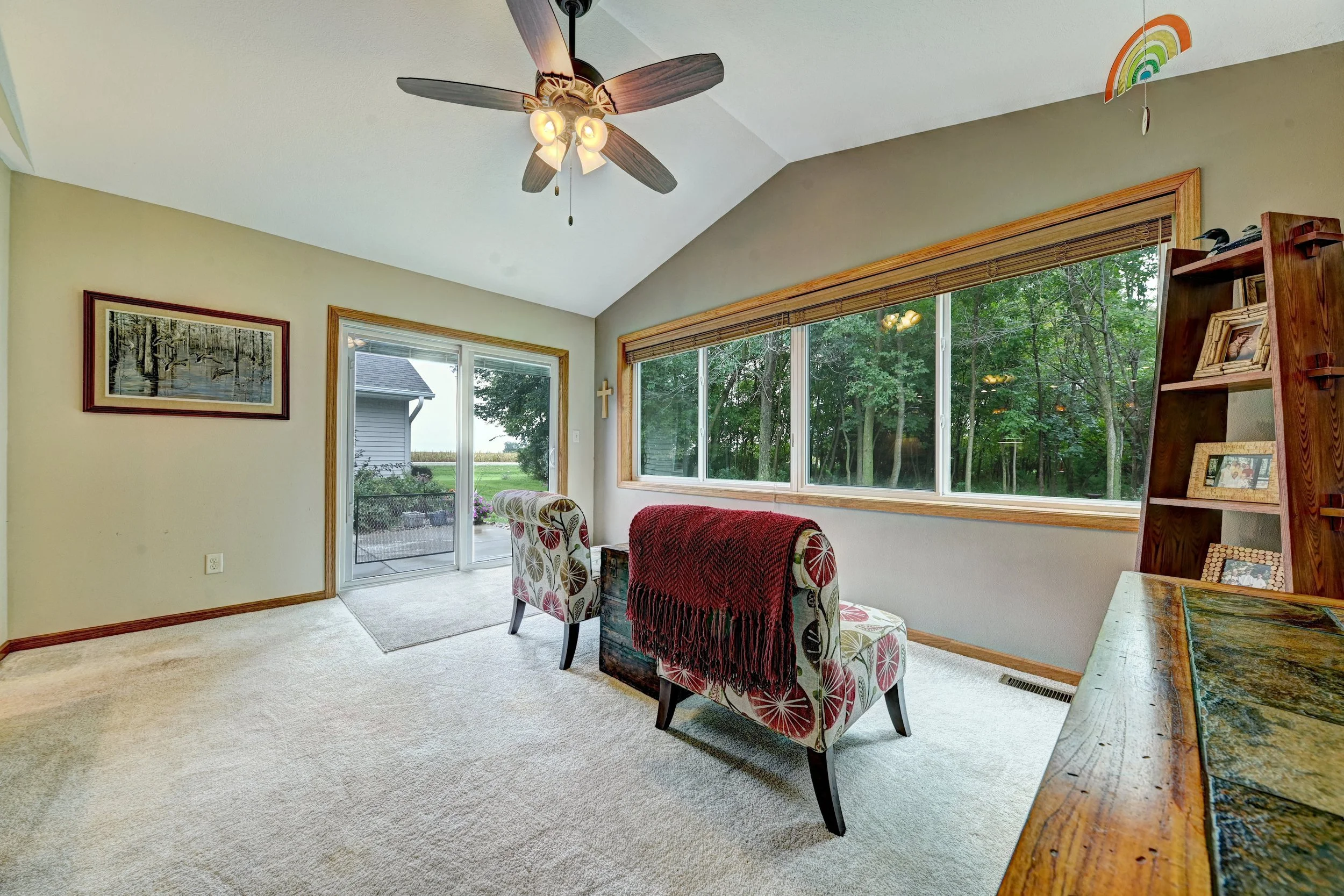 Living room with window view of trees outside, ceiling fan with light, framed artwork on beige wall, armchairs with floral pattern and red throw blankets, wooden shelf with pictures and decorations, and a sliding glass door leading outside.