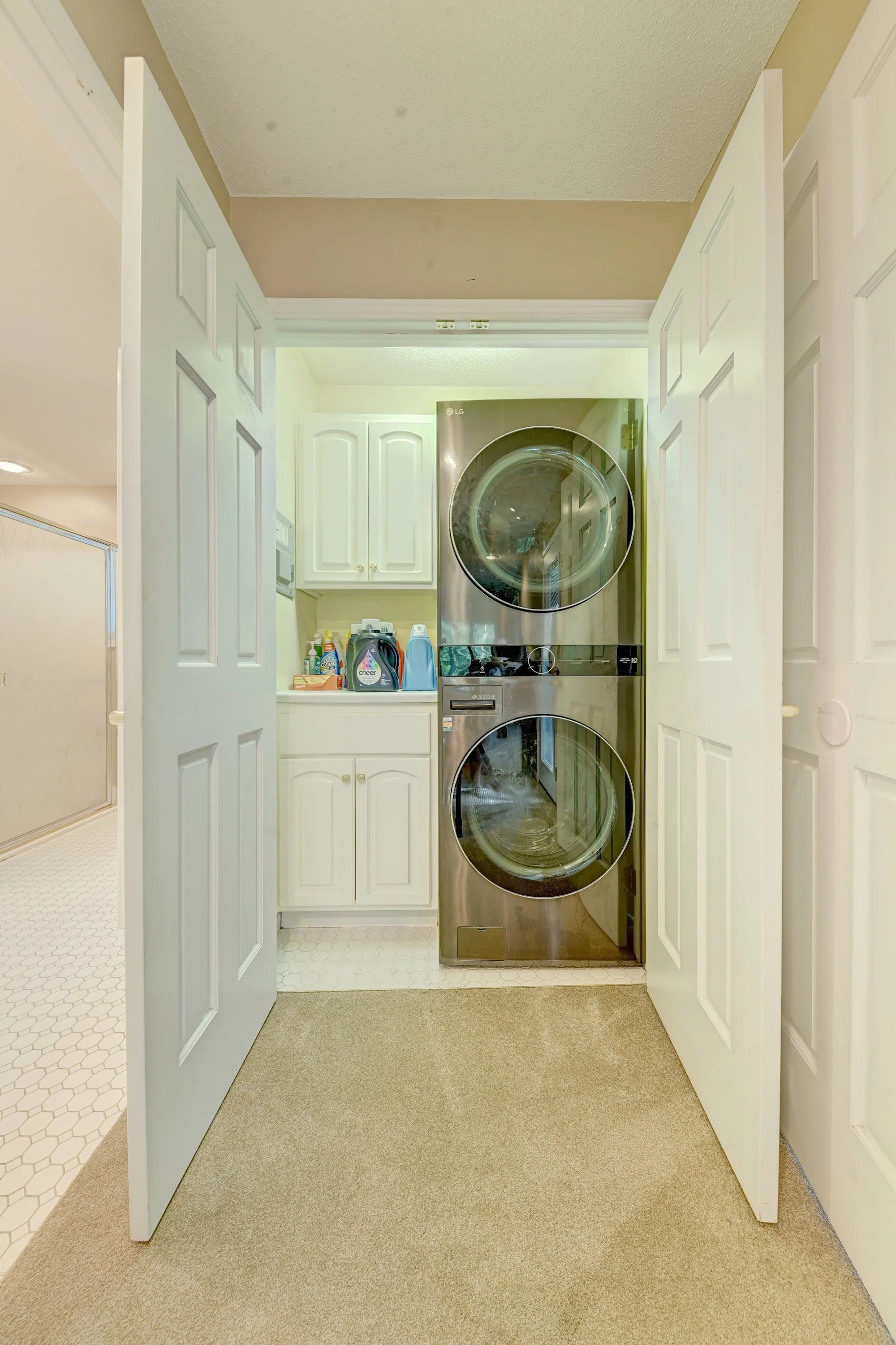 Laundry room with a stainless steel front-loading washer and dryer stacked, white cabinets, and laundry detergents on the counter, viewed through double white doors.