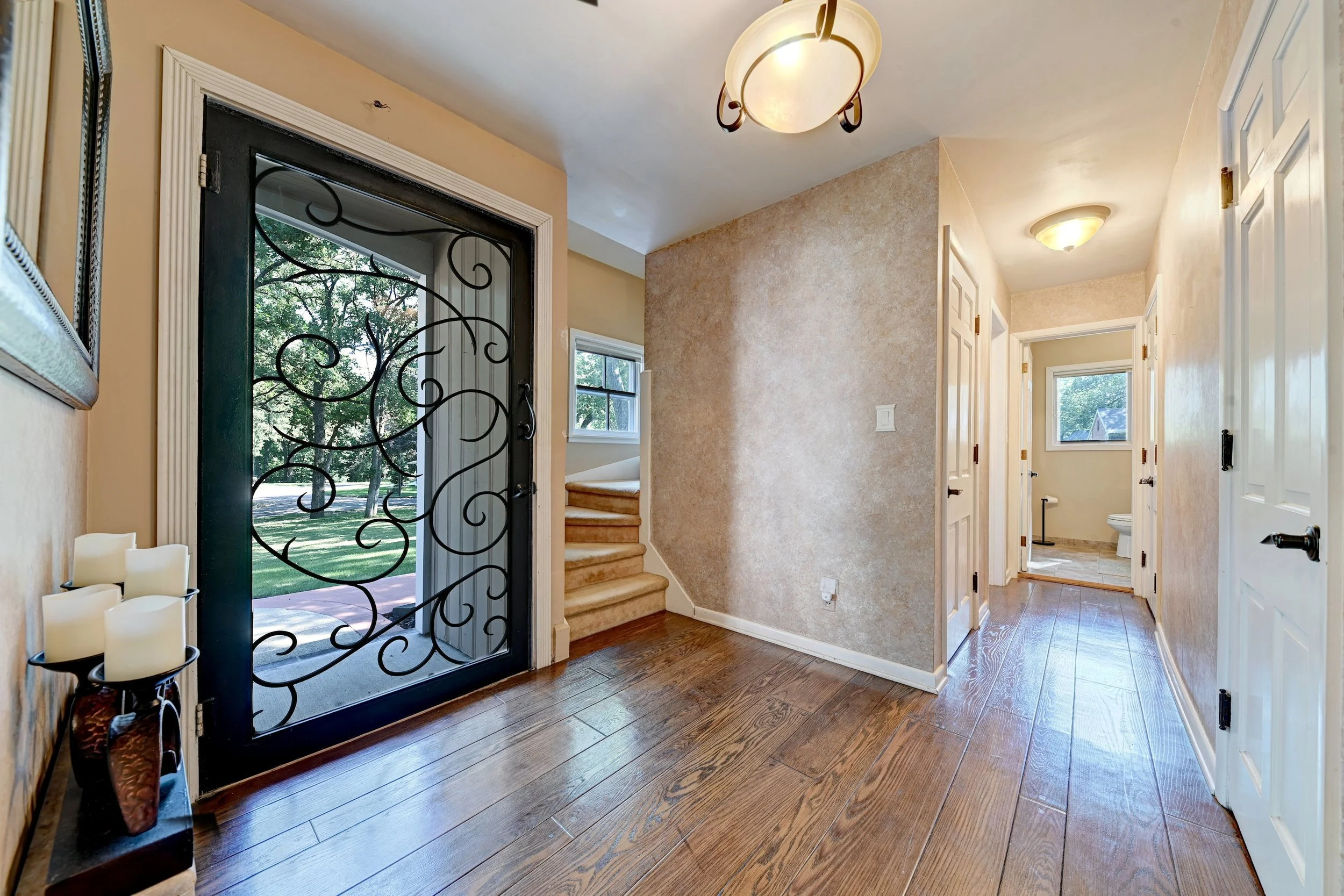 Interior view of a hallway with wood flooring, white door frames, a staircase with beige carpet, and a bathroom with a window and tile flooring at the end.
