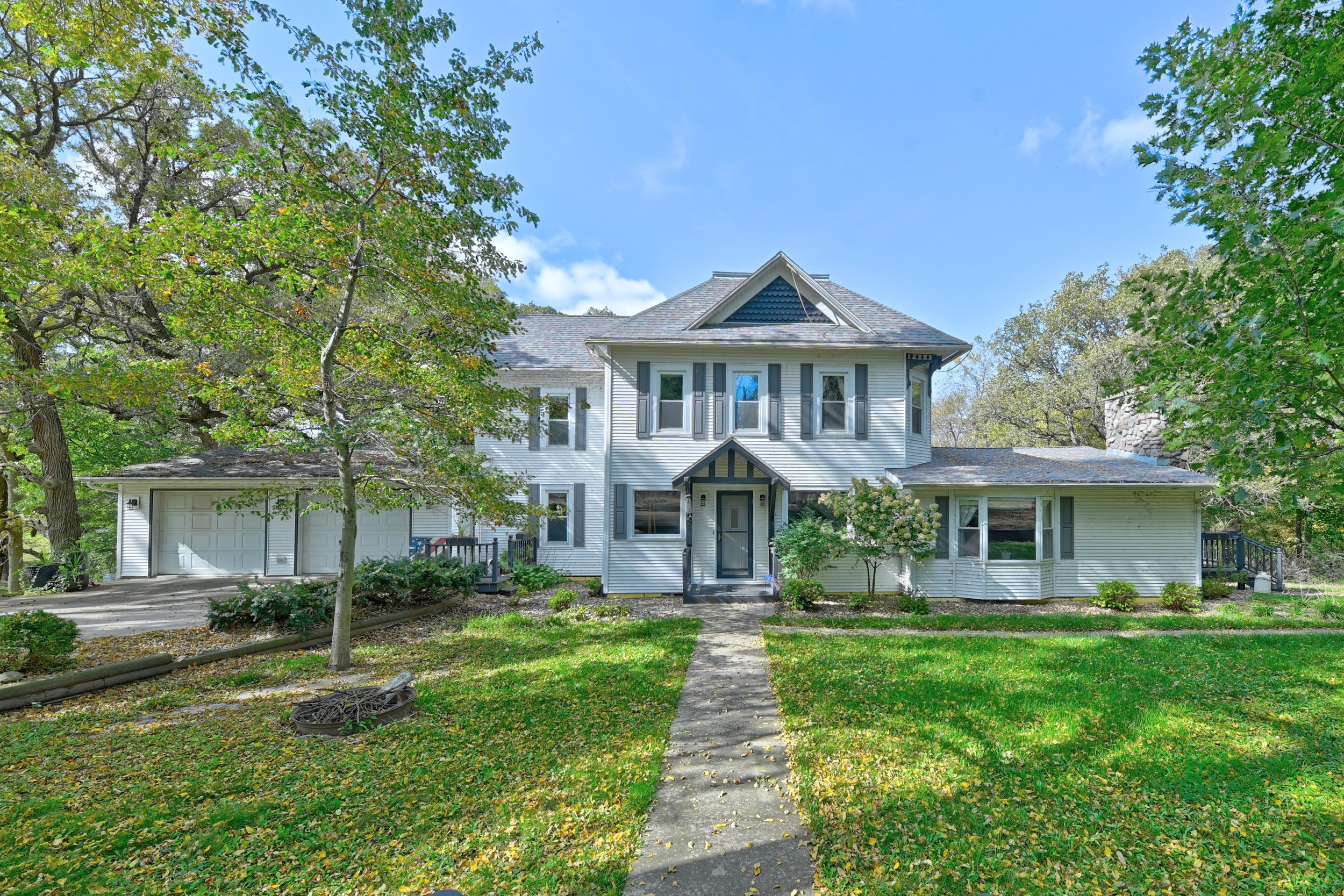 Front view of a two-story white house with a steep roof, surrounded by trees and greenery, under a blue sky.