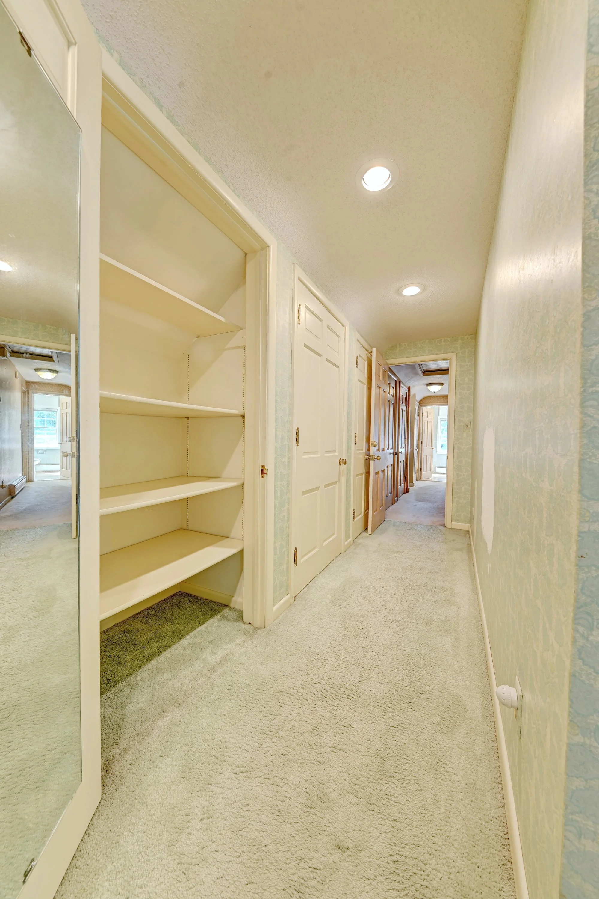 A hallway with beige carpet, greenish patterned wallpaper, white doors, and a closet with sliding door. Recessed ceiling lights and a large mirror on the left wall. Various rooms are visible at the end of the hallway.