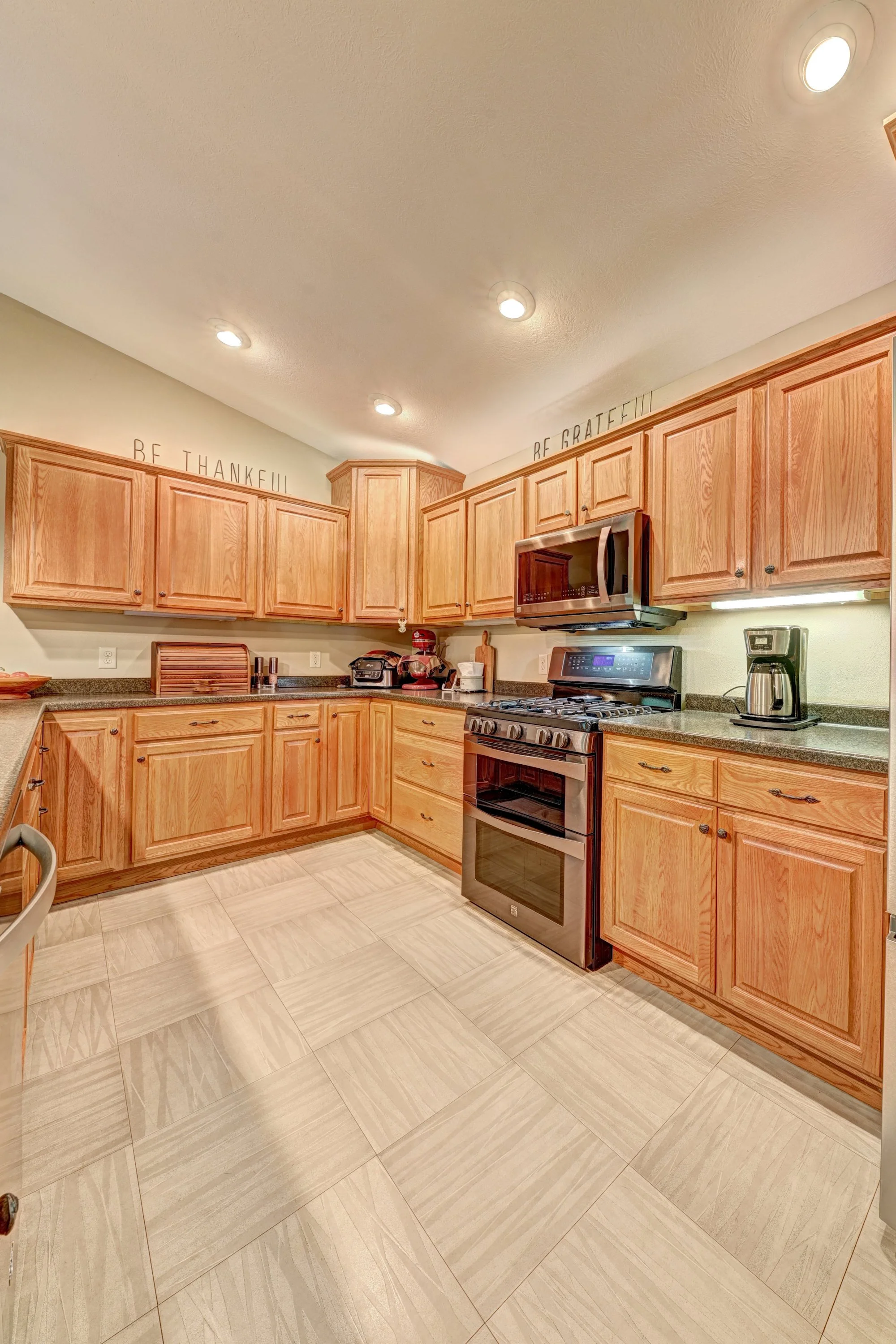 Kitchen with wooden cabinets, a stainless steel oven, microwave, coffee maker, and various small appliances on the countertop. Ceiling lights and wall decorations. Floor tiled in beige.