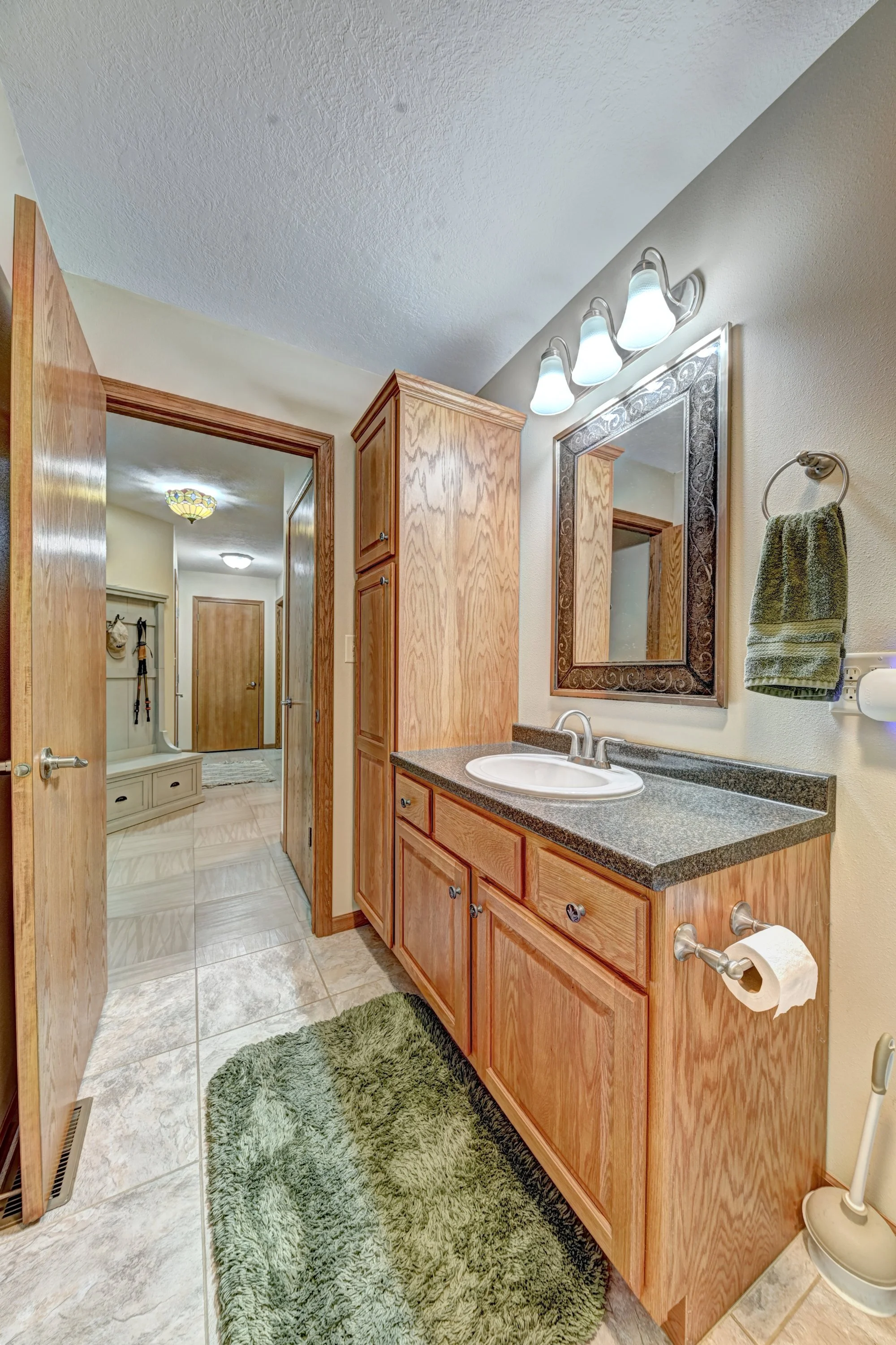Bathroom with wooden cabinet, granite countertop, mirror, wall-mounted light fixture, sink, green shaggy rug, towel on towel ring, toilet paper holder.