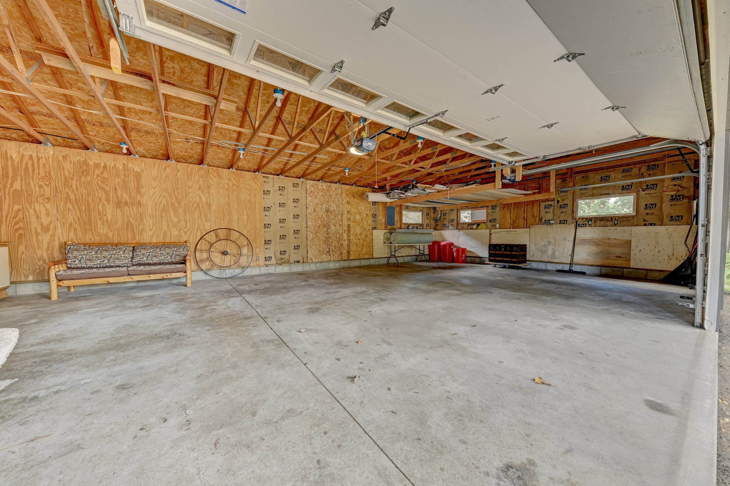 Empty garage with partly opened garage door, unfinished walls with insulation, a couch with a geometric pattern, some red containers, and construction tools and materials.