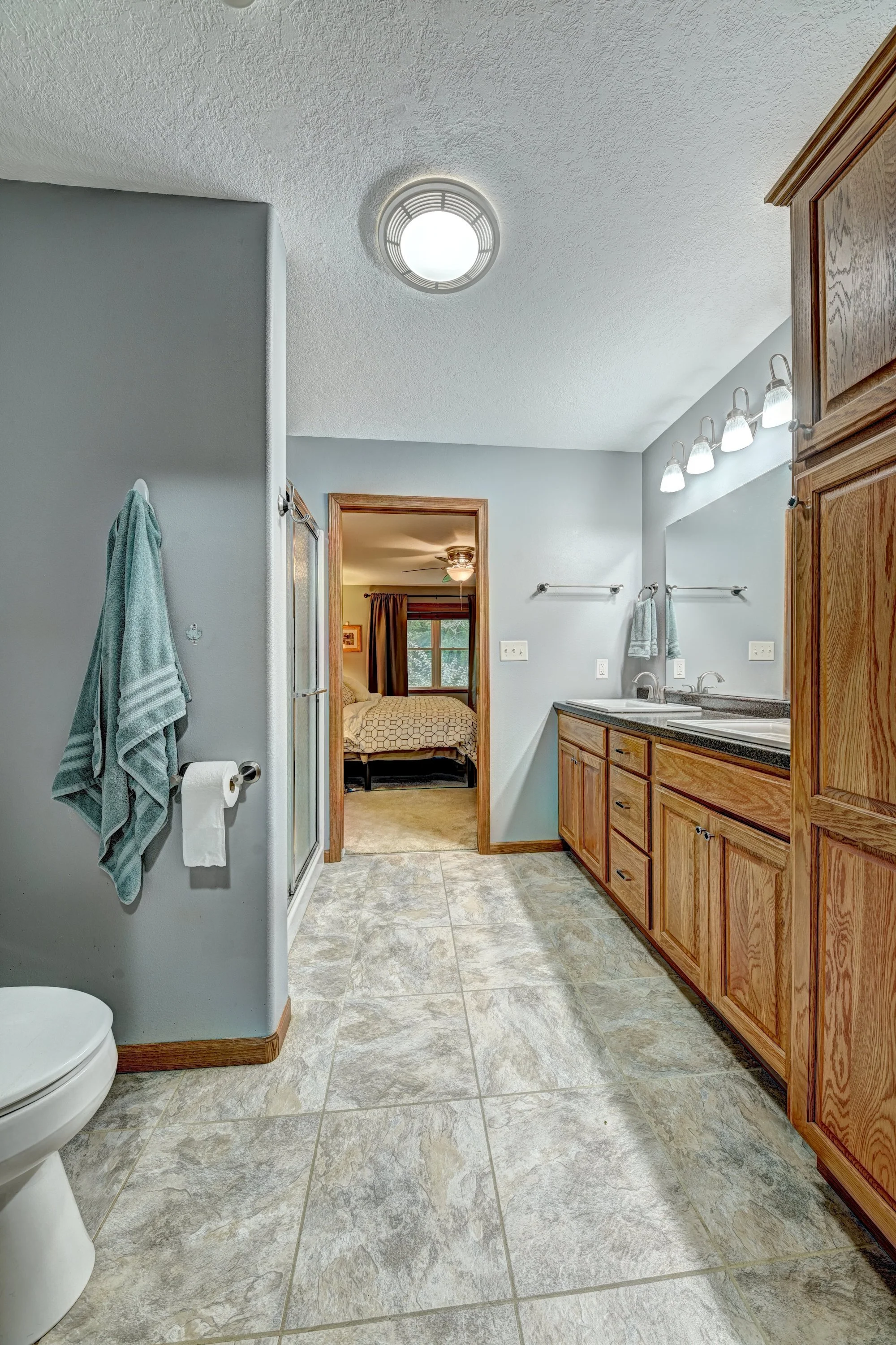 The image shows a bathroom with a shower stall on the left, hanging towels, a toilet in the corner, a wooden cabinet on the right, a sink and mirror with lights above, and a view into a bedroom through a doorway. The bathroom has gray walls and tiled