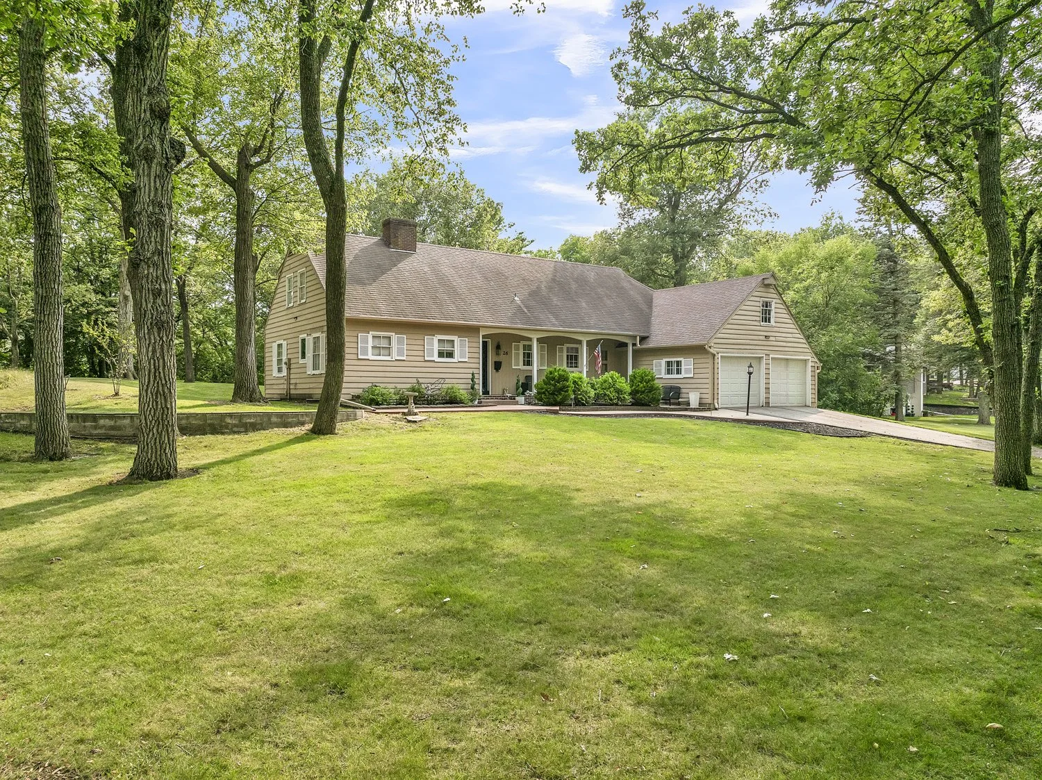 A beige house with a brown roof, surrounded by green trees and a well-maintained lawn, under a partly cloudy sky.
