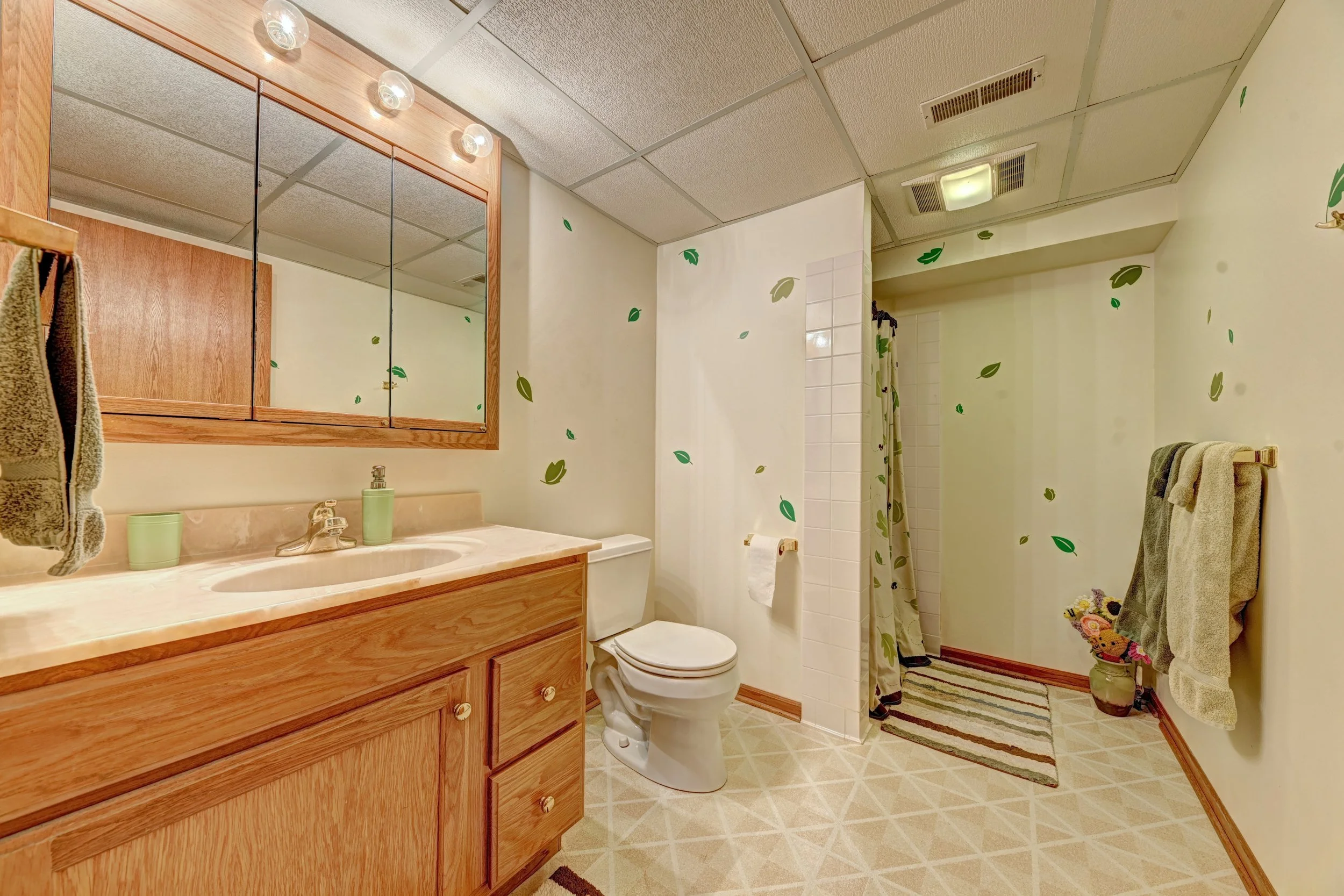 A bathroom with a wooden vanity, a mirror cabinet, a toilet, and a shower with a curtain decorated with green leaves. There are towels hanging on the towel racks and a small rug near the shower. The ceiling has a vent and a light fixture.