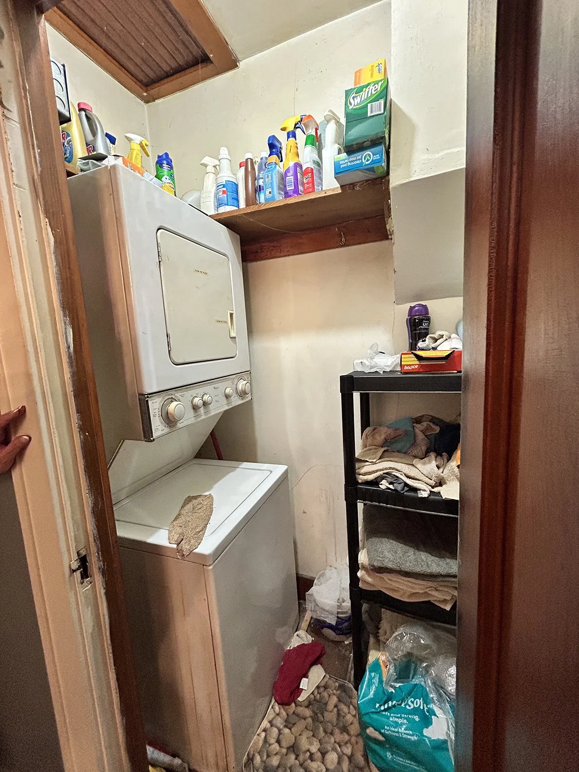 A small laundry room with a stacked washer and dryer, shelves with cleaning supplies, a black storage cart with towels, and various laundry items on the floor.