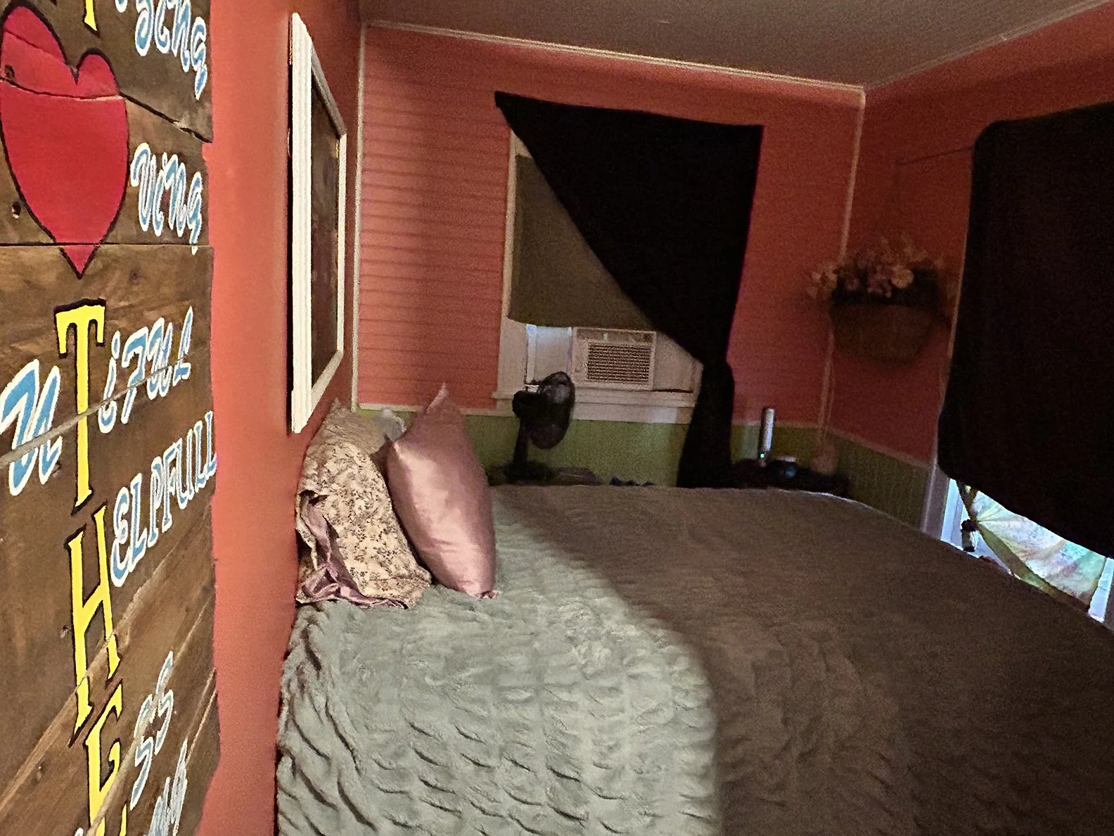 A bedroom with pink and green walls, a bed with two pillows, a wall-mounted air conditioning unit, a window with dark curtains, and a small fan on a nightstand.