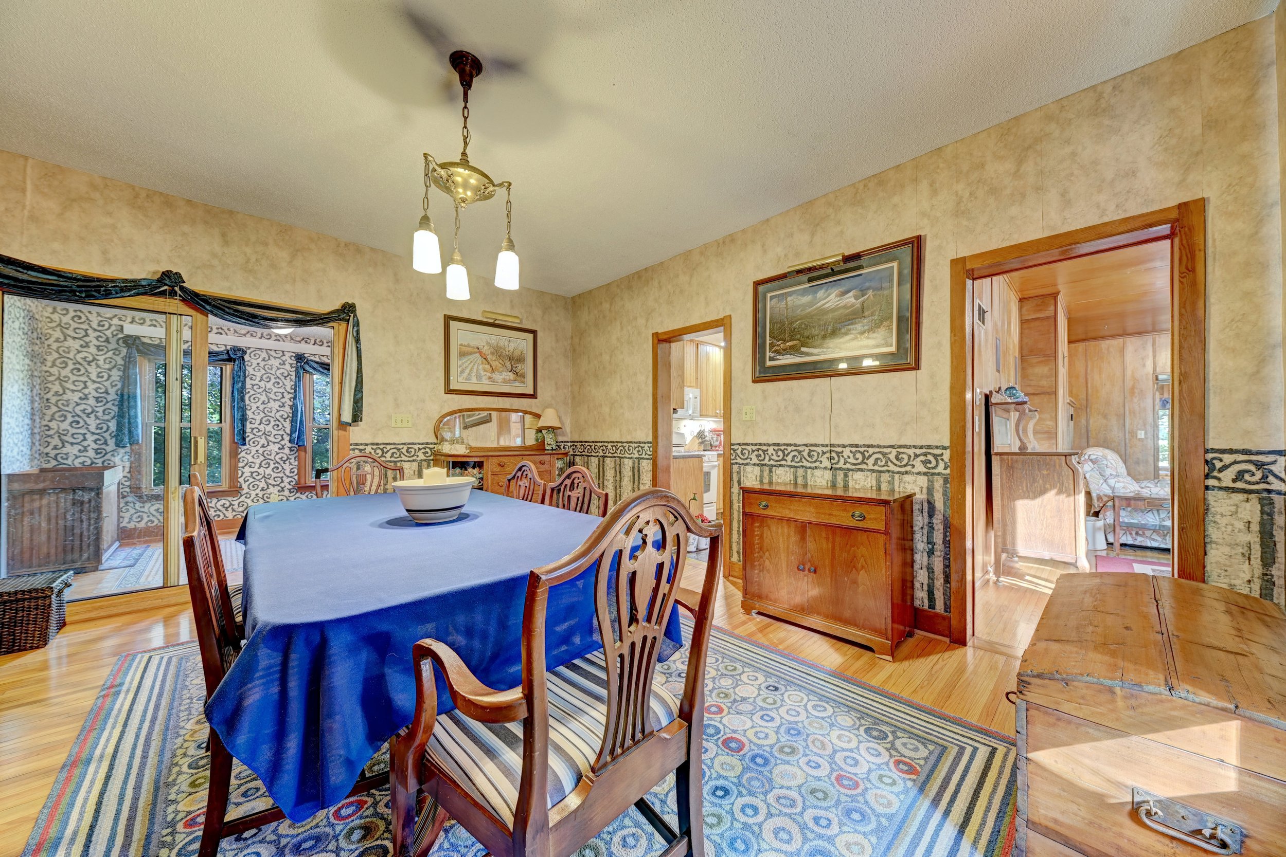 A dining room with a wooden table covered by a blue tablecloth, surrounded by wooden chairs with striped cushions. The room has beige patterned wallpaper, two framed landscape paintings, a window with decorative curtains, a wooden cabinet, a wooden d