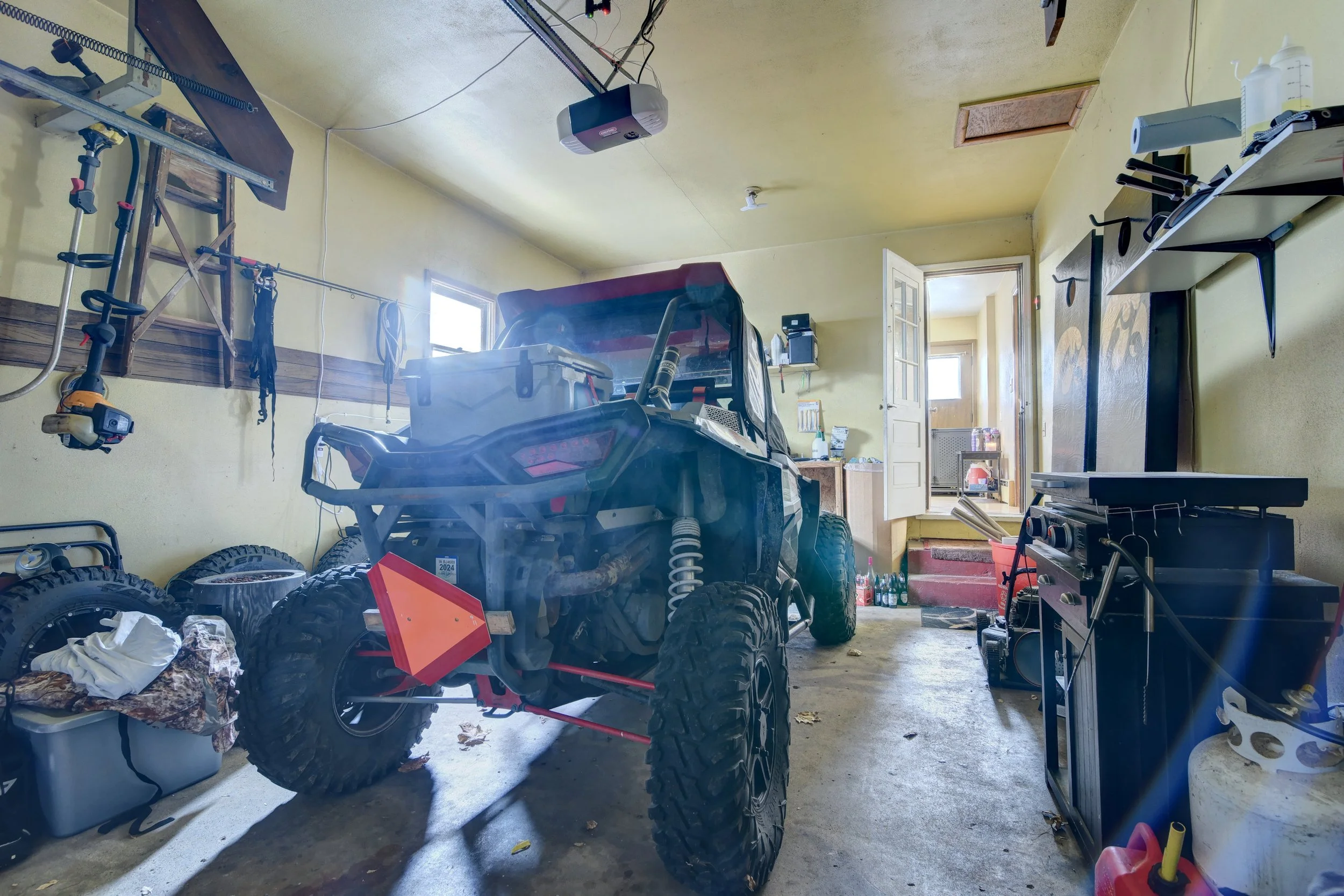 A garage with a dirt bike in the center, tires stacked on the left, and various tools and supplies on shelves and workbenches.