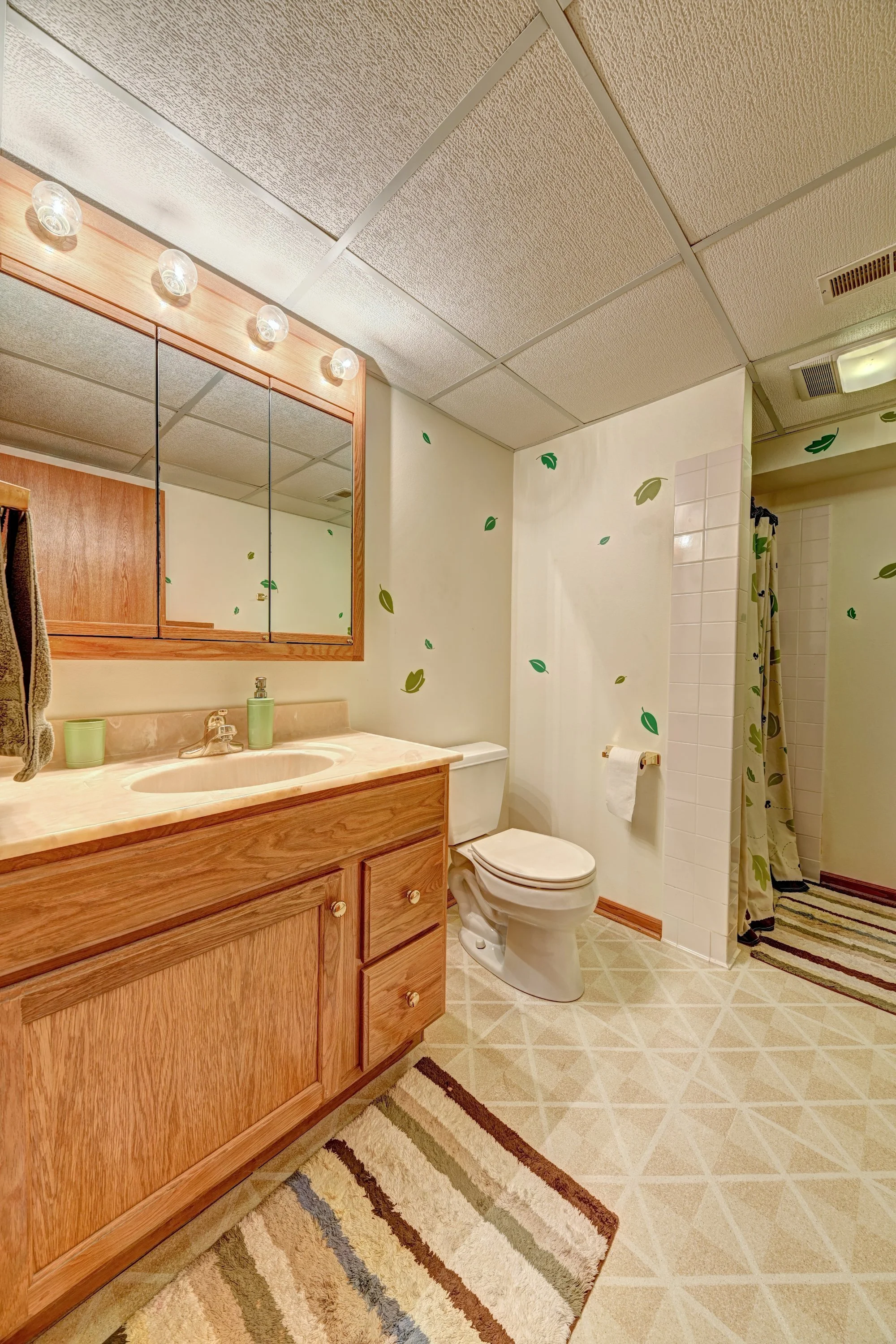 A bathroom with a wooden vanity, mirror cabinet, toilet, shower with a curtain, and a striped rug on tile flooring.