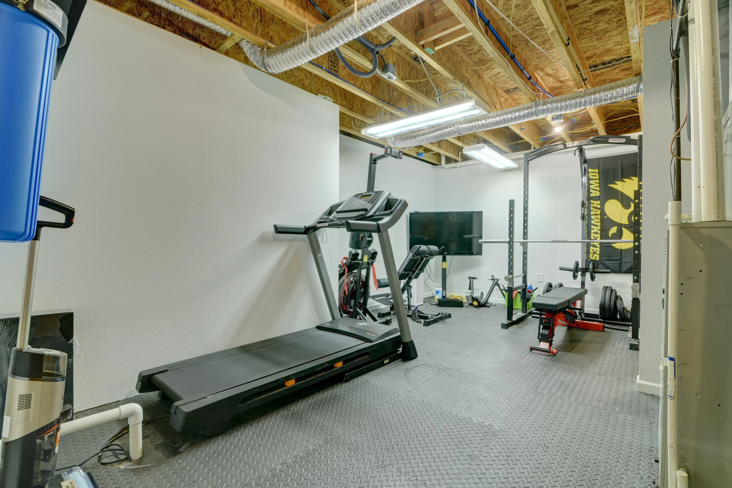 A small home gym with gym equipment including a treadmill, squat rack, weight bench, and weights, with a television and unfinished wooden ceiling.