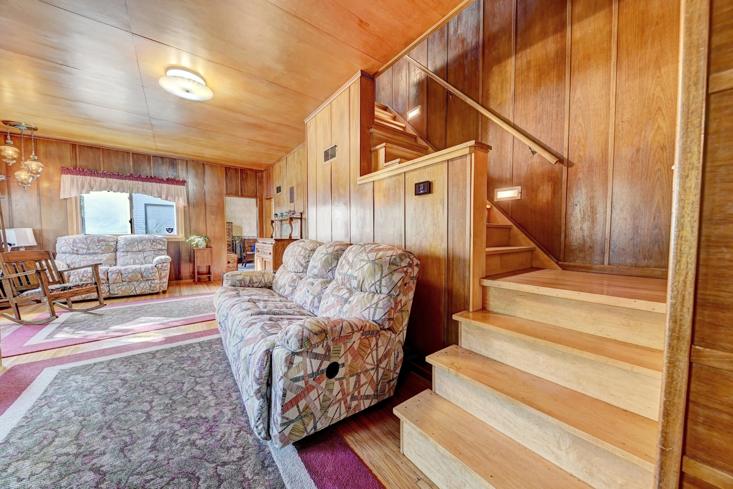 Living room with wood-paneled walls and ceiling, floral-patterned sofas, a wooden staircase, and a window with pink and white curtains.