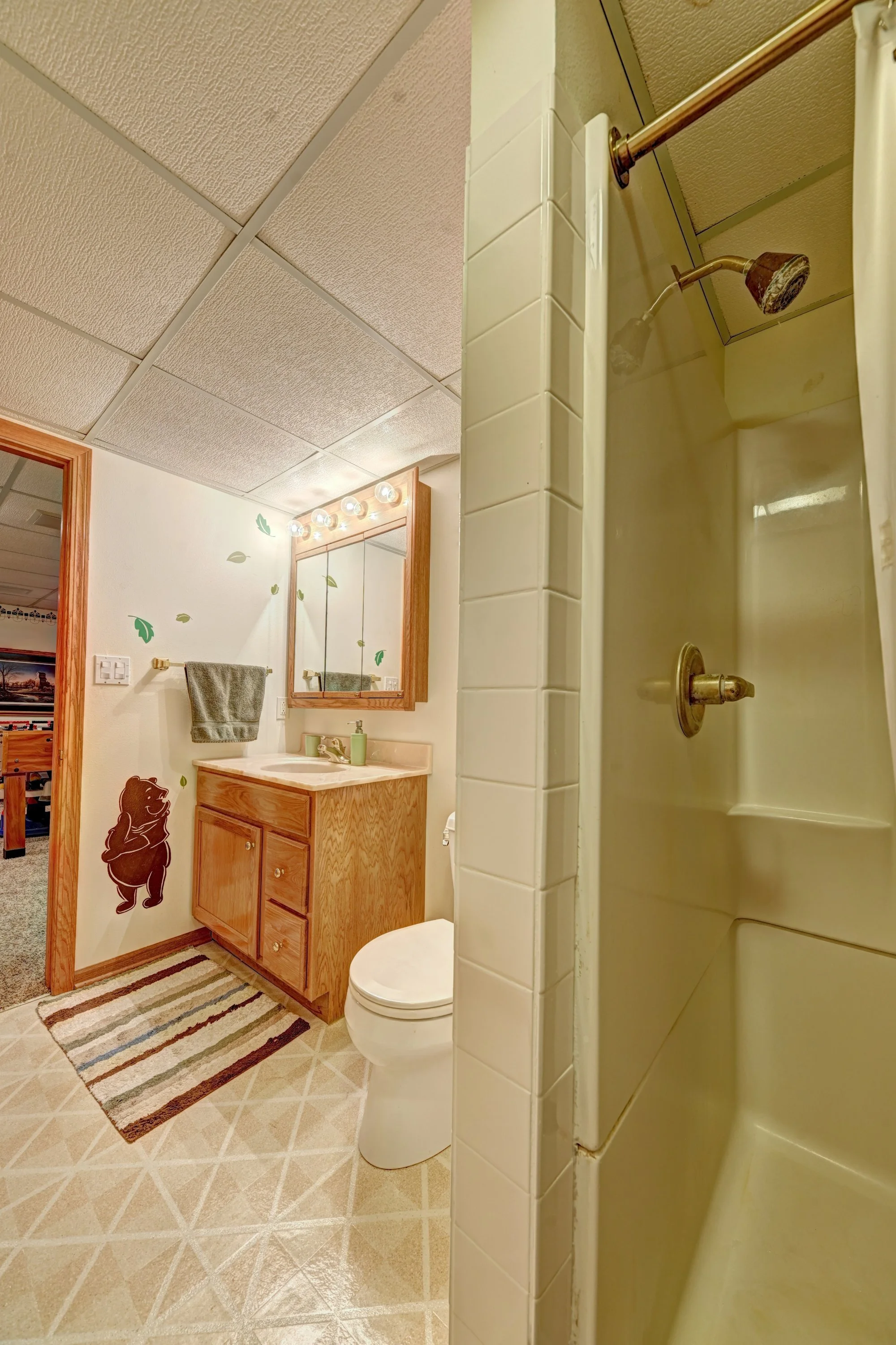 Bathroom with a wooden framed mirror, a vanity with a sink, a towel hanging, a toilet, and a shower with brass fixtures. The walls are decorated with bear stickers and the floor has a striped rug.