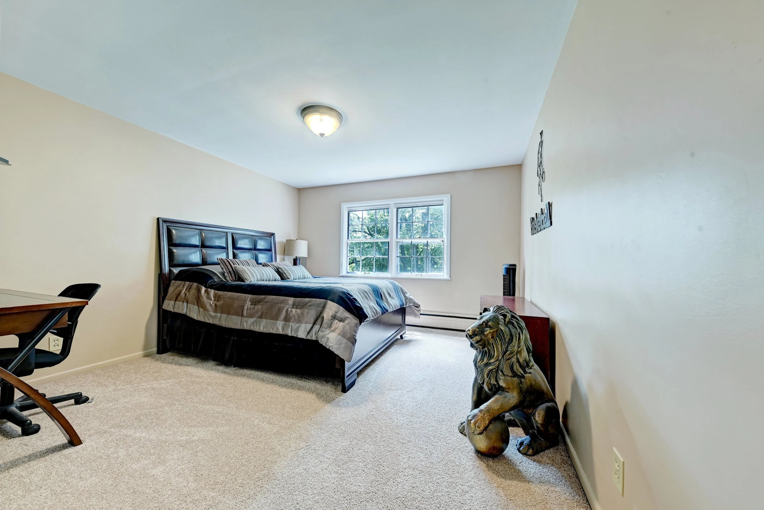 A bedroom with beige walls, a window with trees outside, a bed with striped bedding, a wooden nightstand with a lamp, a decorated statue of a lion, and a side table.