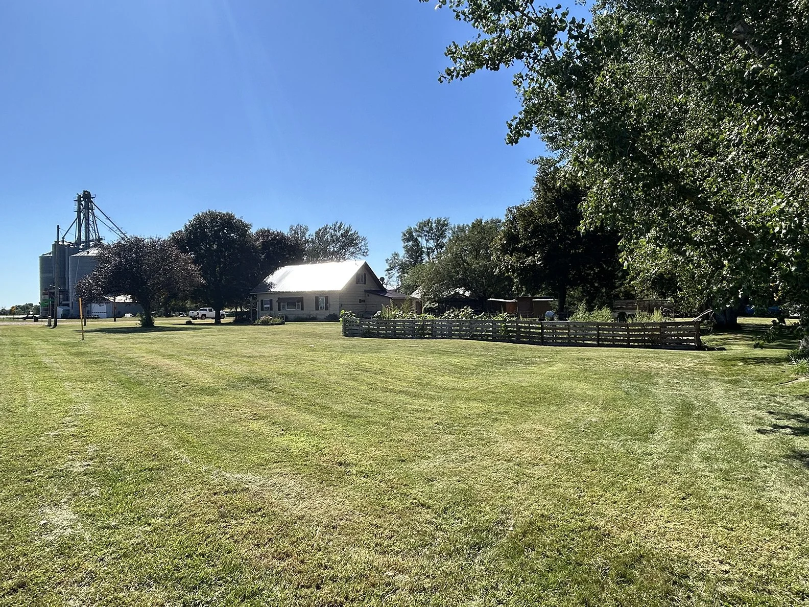 A rural farmland scene with a well-maintained grassy field, a white farmhouse, trees, a wooden fence, and silos under a clear blue sky.