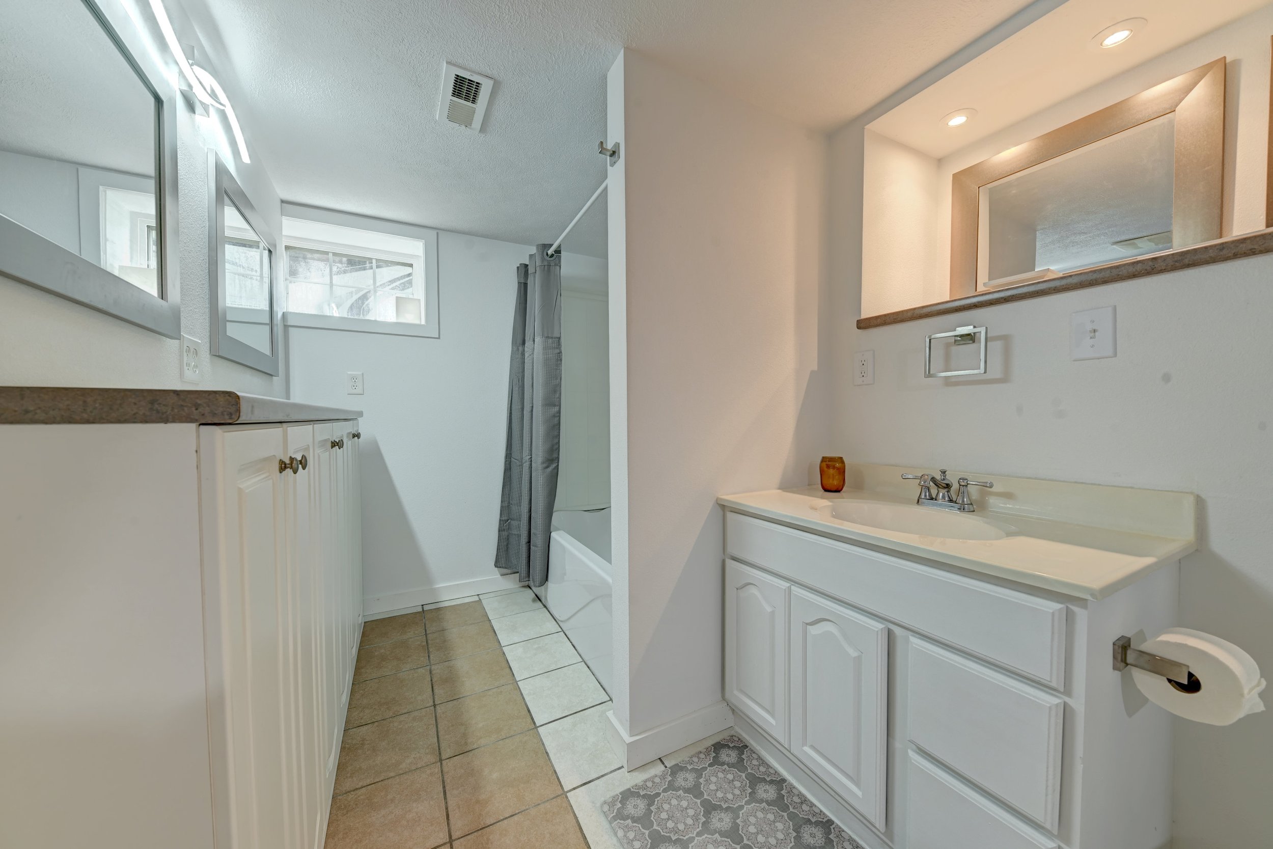 Bathroom with white cabinets, beige tiled floor, and a sink with a mirror above it. A window and shower with a gray curtain are visible in the background. There are small decorative items and a soap dispenser near the sink.