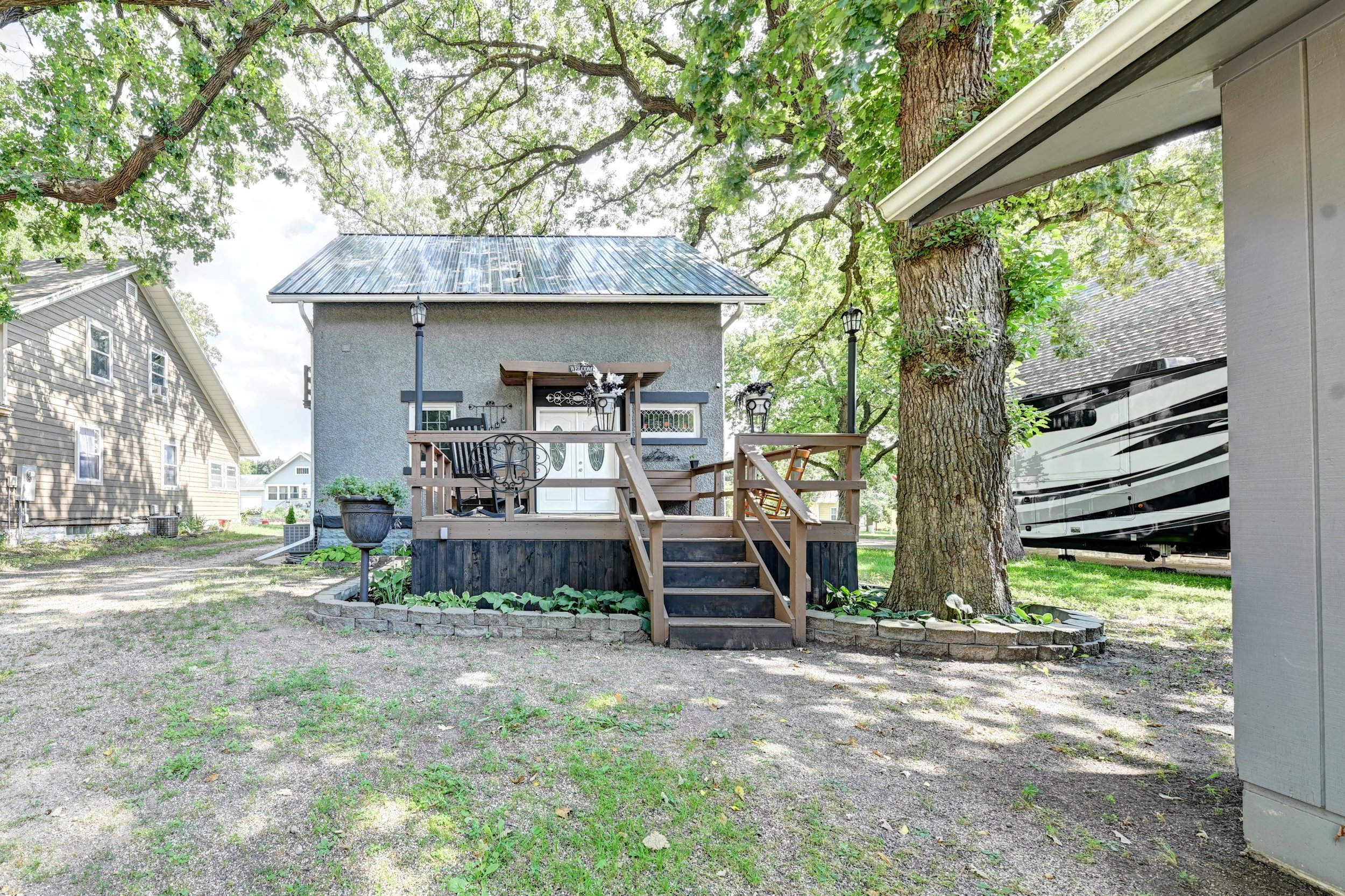 A backyard scene with a small gray house on a wooden deck, surrounded by green trees and grass. There are two black streetlamp-style lights on the deck, and a large tree with a textured trunk in the foreground. To the right, part of a house and a lar